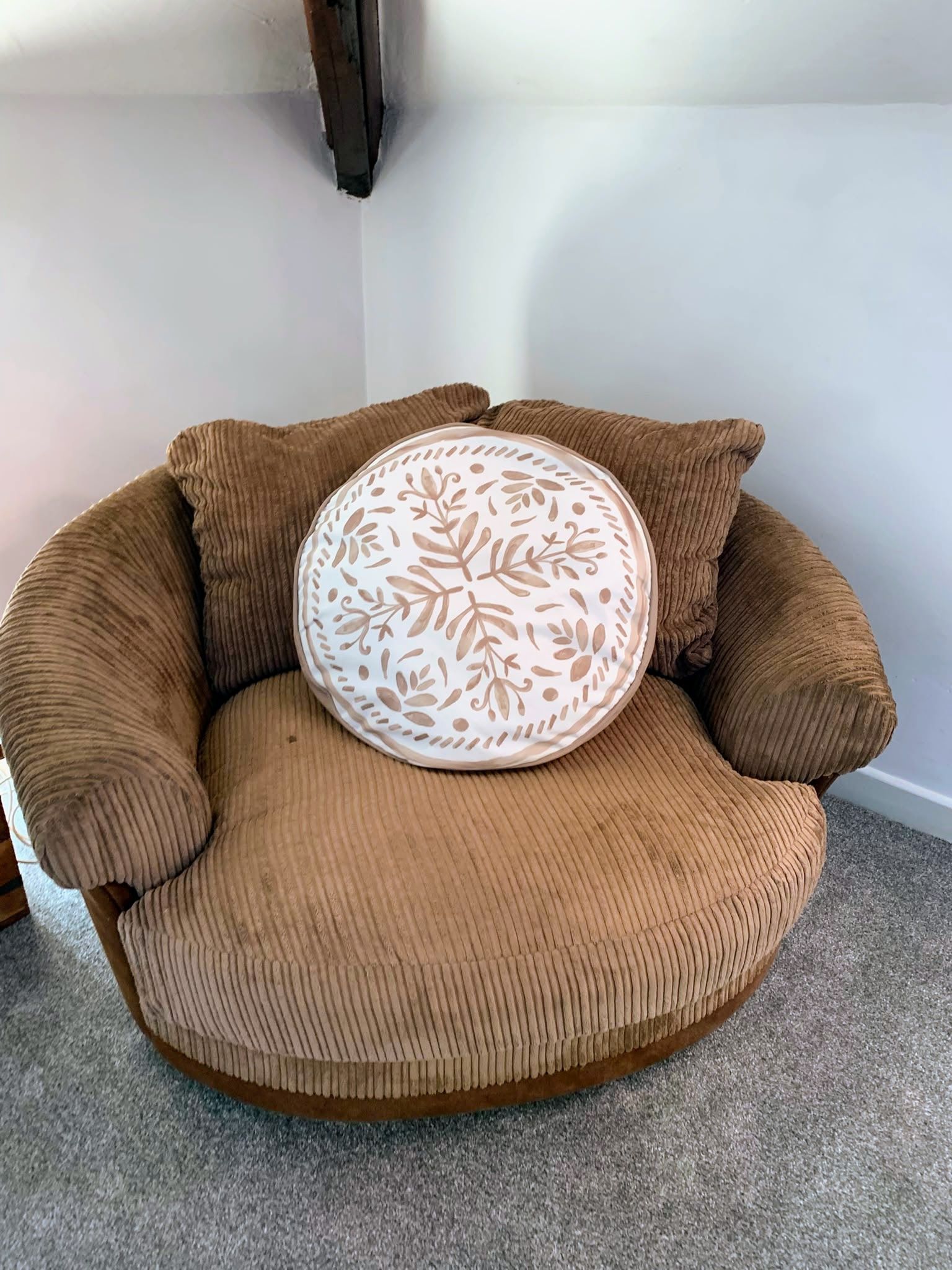 Brown upholstered chair with a round white patterned pillow in a corner against a white wall