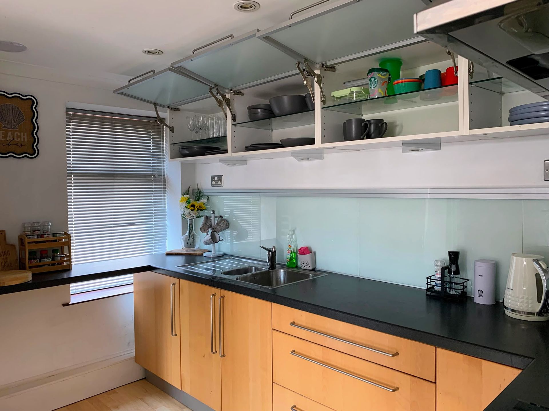 Modern kitchen with black countertops, light wood cabinets, sink, shelves, and a window with blinds