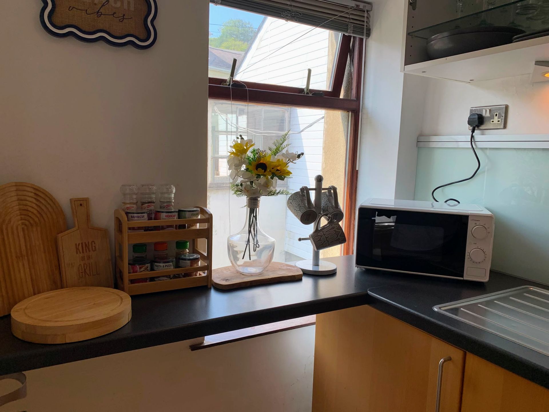Kitchen counter with a vase of flowers, microwave, sink, and window with blinds open