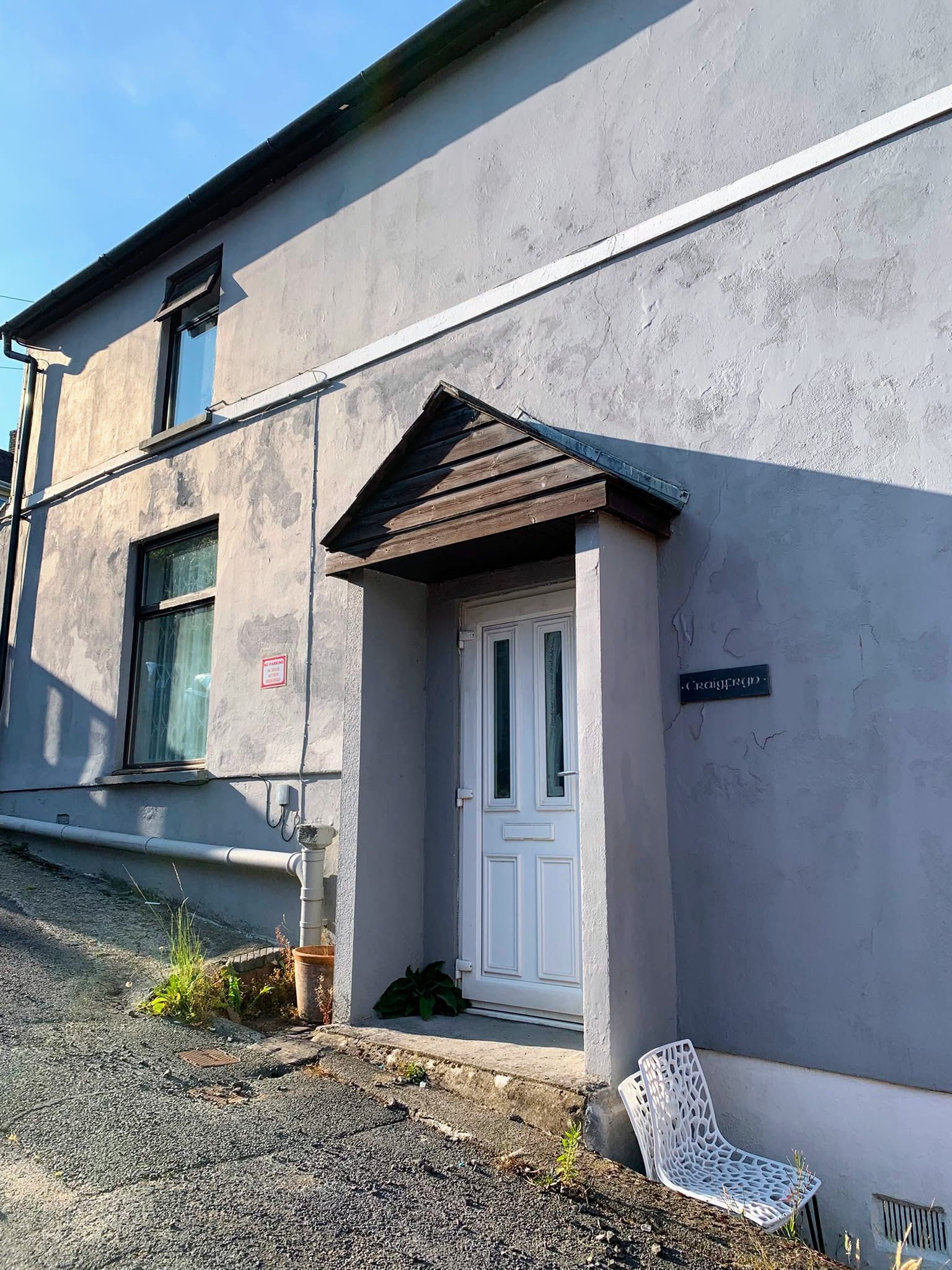 Small gray house exterior with a white front door and a tiny awning over a gravel yard.