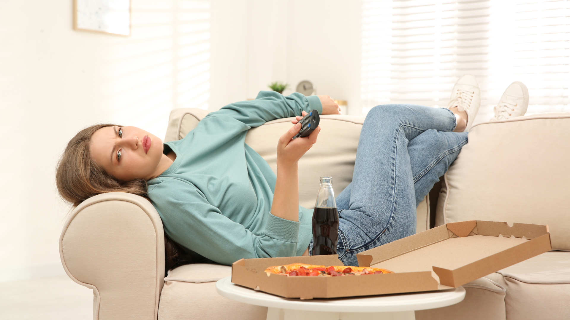 A woman is laying on a couch with a box of pizza and a remote control.