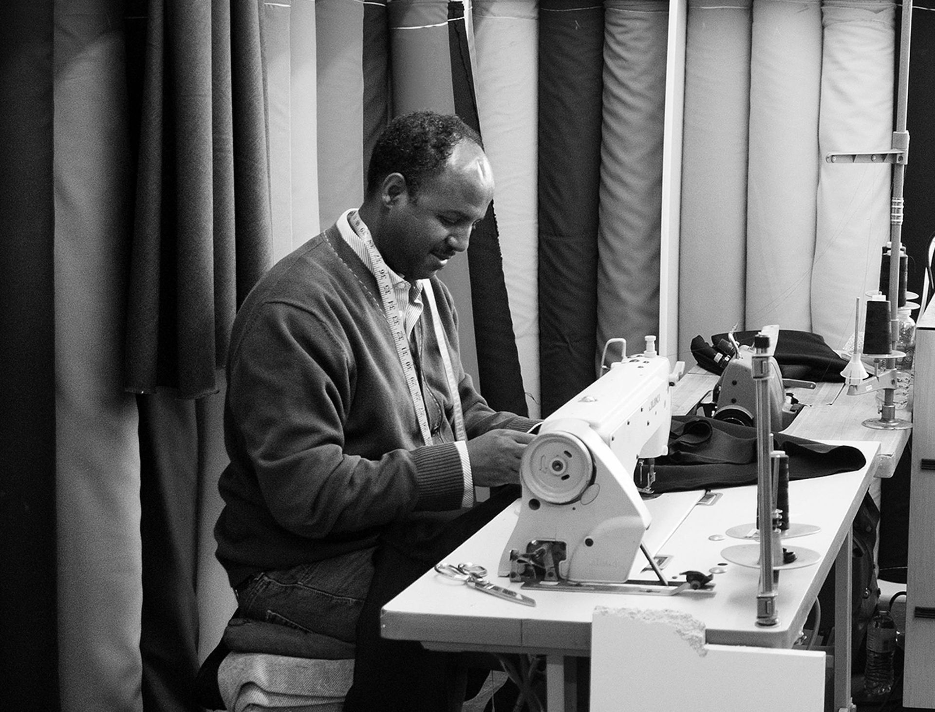 Black and white photo of a man sewing at a machine. Fabric rolls are in the background.