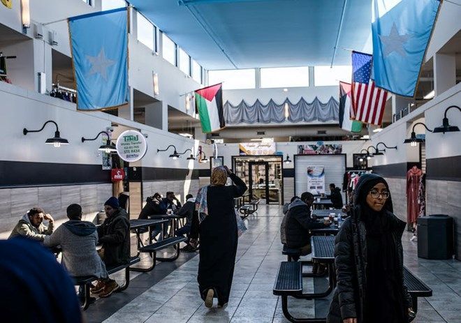 People in a food court with flags hanging, including Somalia and US. Several people are dining at tables.