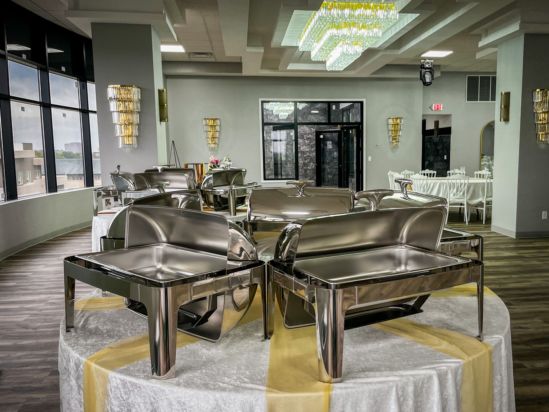 Buffet setup in an event space, featuring stainless steel chafing dishes on a round table with gold accents.