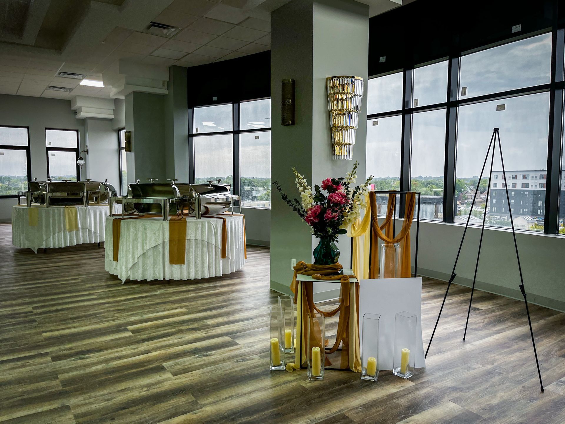 Event space with buffet tables, floral arrangement, and a sign on an easel, all near large windows.