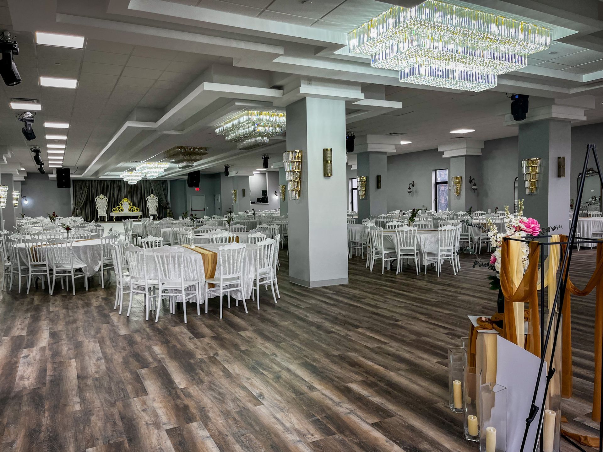 Spacious banquet hall with white tables and chairs, chandeliers, and wood floors.