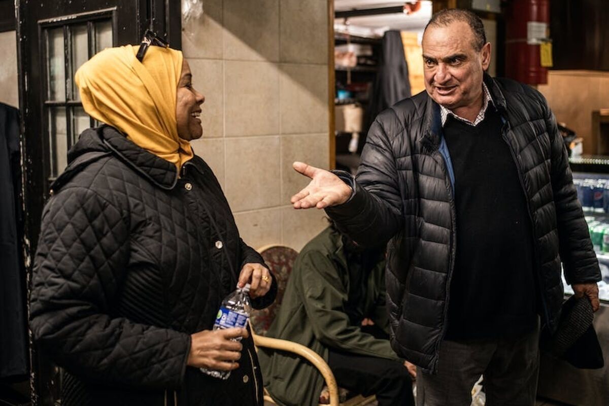 Woman in yellow hijab chats with man in black jacket; inside doorway.