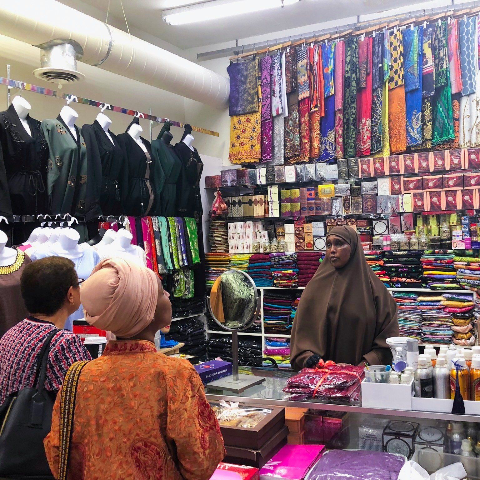 People shopping at a clothing store with headscarves and dresses, a woman behind the counter, and colorful fabrics.