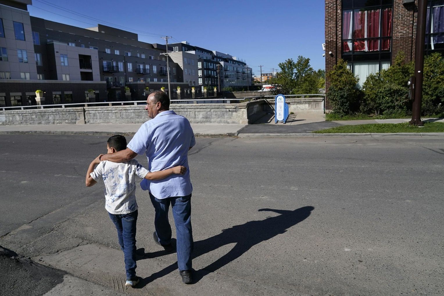 Man with arm around boy walking in street, city buildings in background. Sunny day.