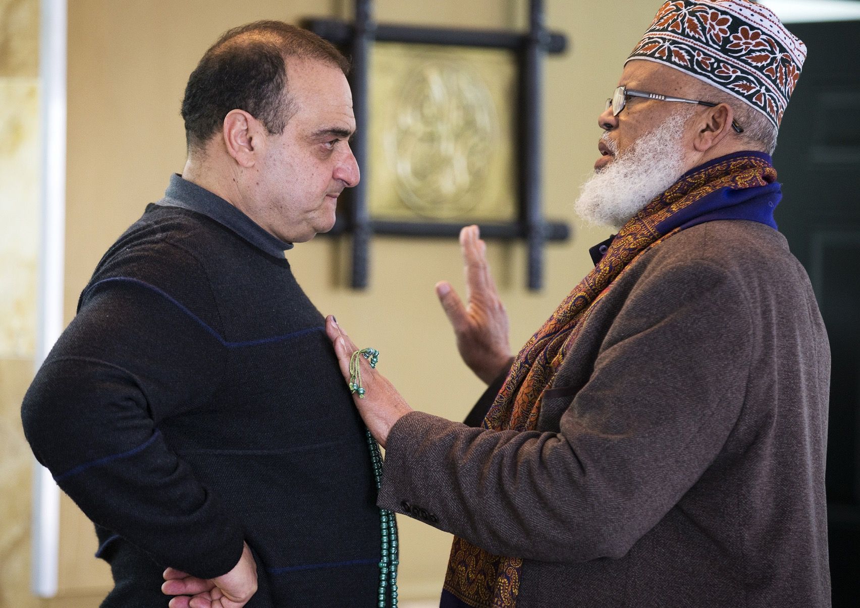 Two men talking; one in a black sweater, the other in a brown coat with a patterned cap and beard.