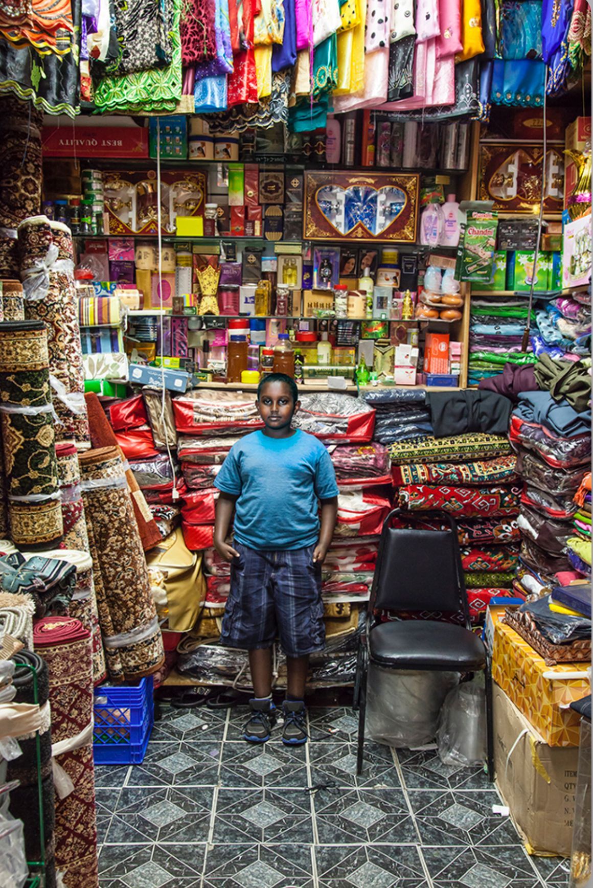 Boy in blue shirt and patterned shorts stands in a cluttered shop filled with products.