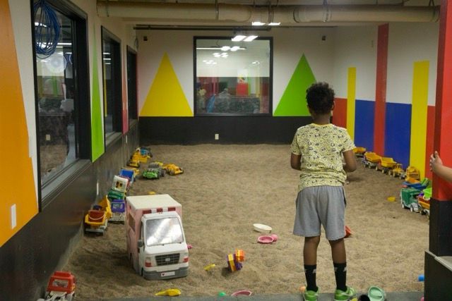 Boy stands in a sandbox with toy cars and trucks. Brightly colored walls and a window are in the background.