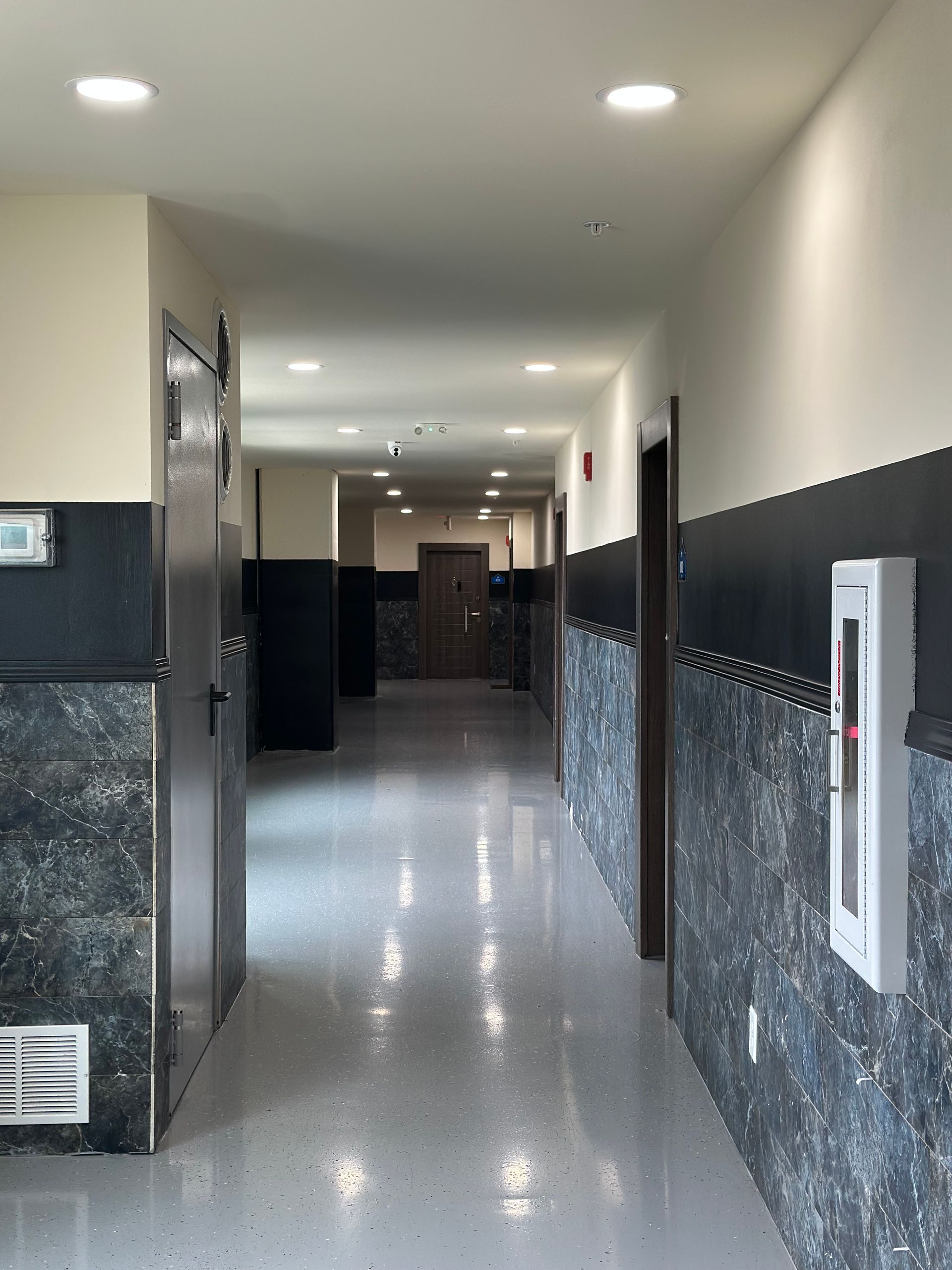 Long, narrow hallway with black and grey panel walls, doors, and a fire extinguisher.