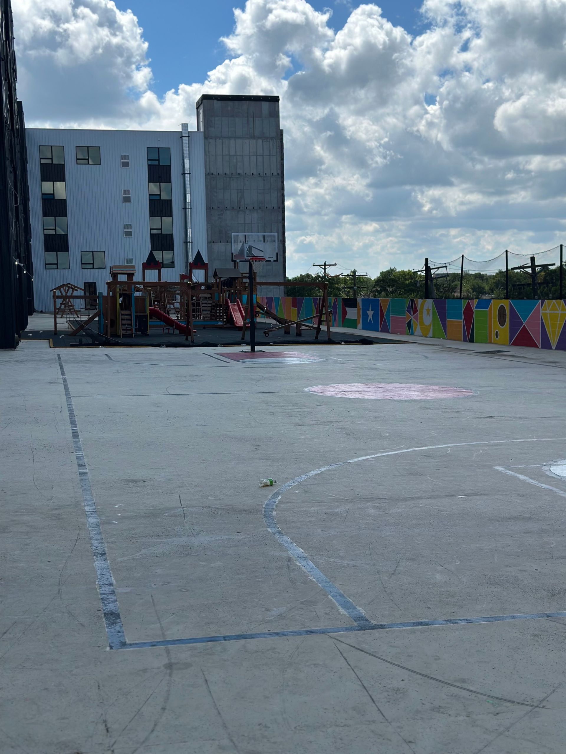 Playground on a rooftop. Basketball court with play structure in the background. Colorful mural along wall.