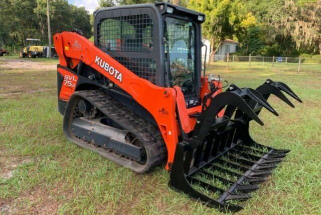 A kubota skid steer with a grapple attached to it is parked in a grassy field.