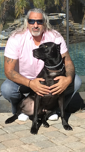 A man is kneeling down next to a black dog in front of a pool.