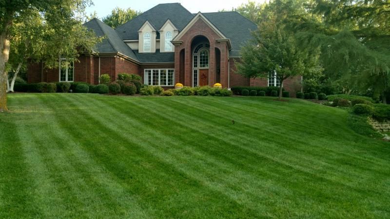 A large, red brick two-story house with a grey roof and neatly mowed lawn in a suburban neighborhood setting.