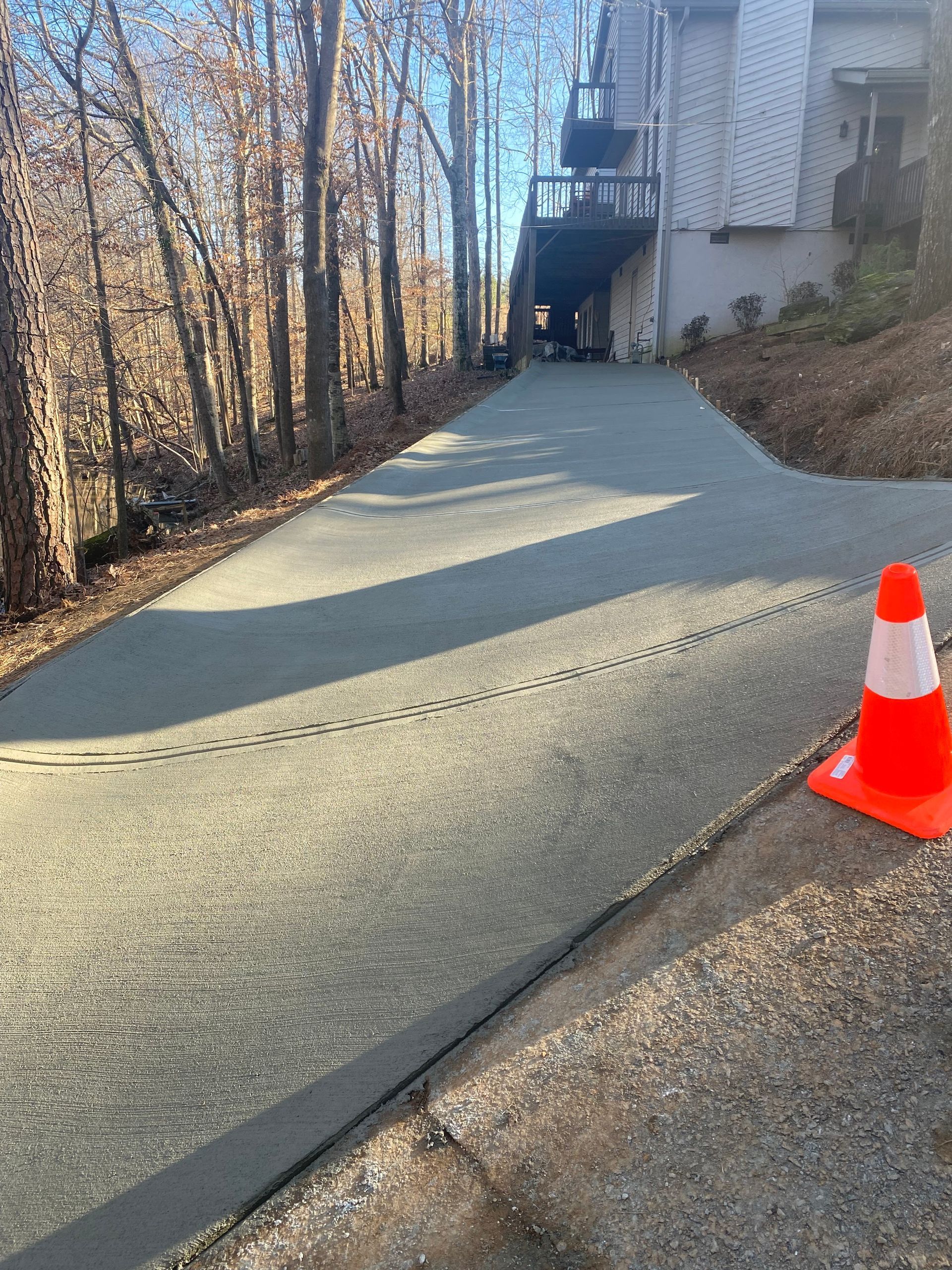 Newly paved concrete driveway leading uphill towards a house. Orange traffic cone on the right.