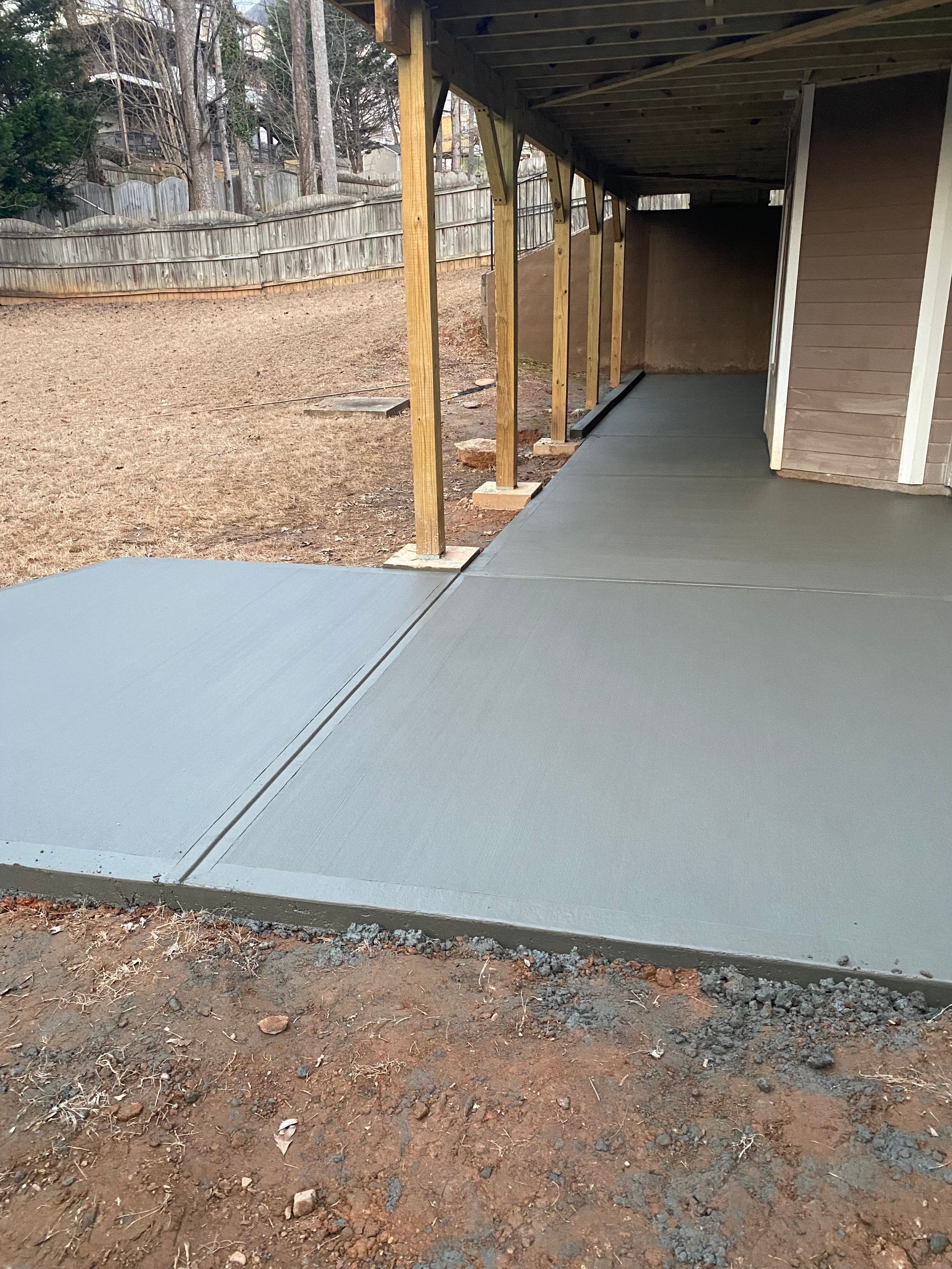 Newly poured gray concrete patio under a wooden porch roof. Dirt surrounds the patio.