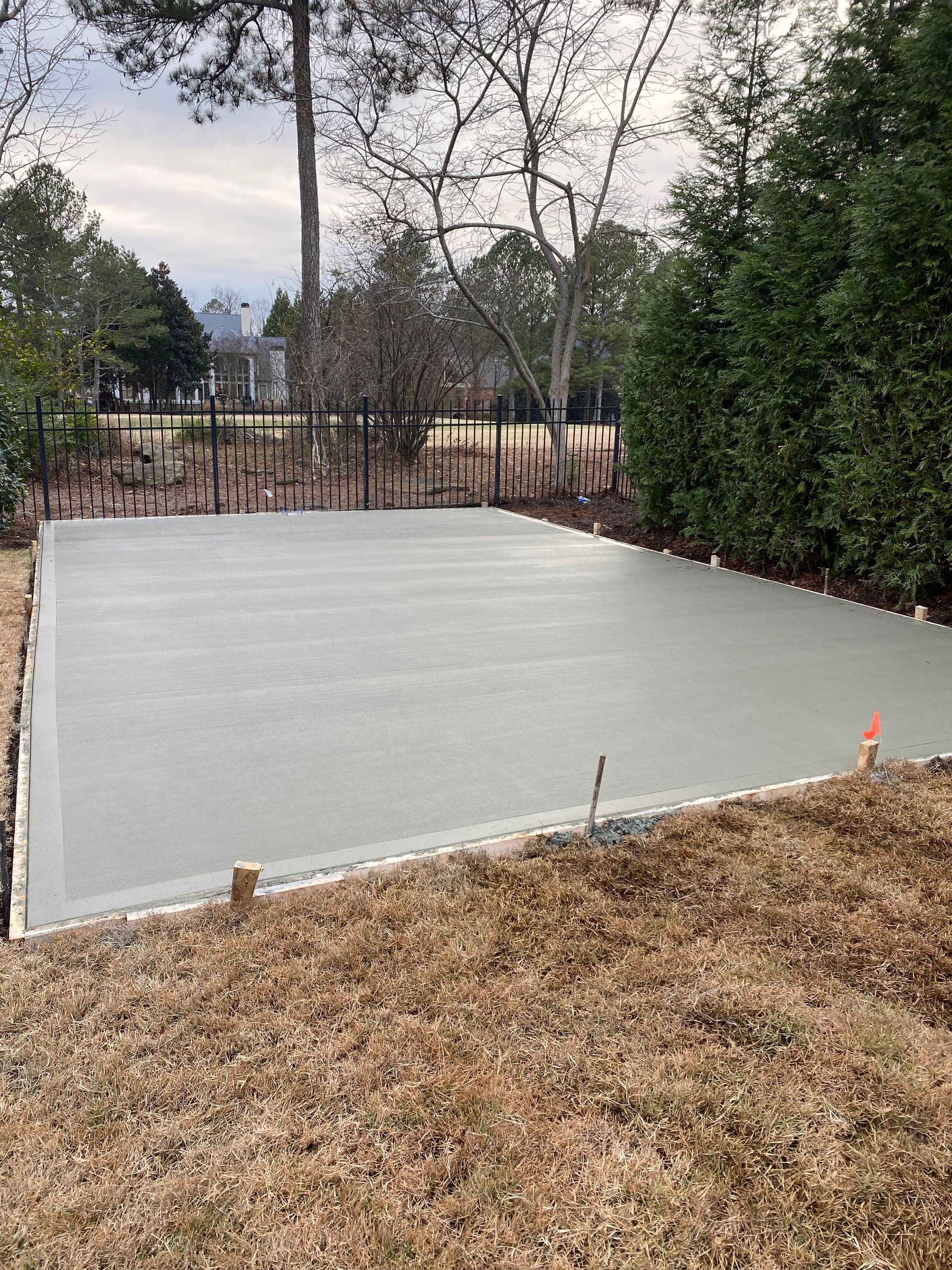 Newly poured concrete slab in a backyard, surrounded by brown grass, trees, and a black fence.