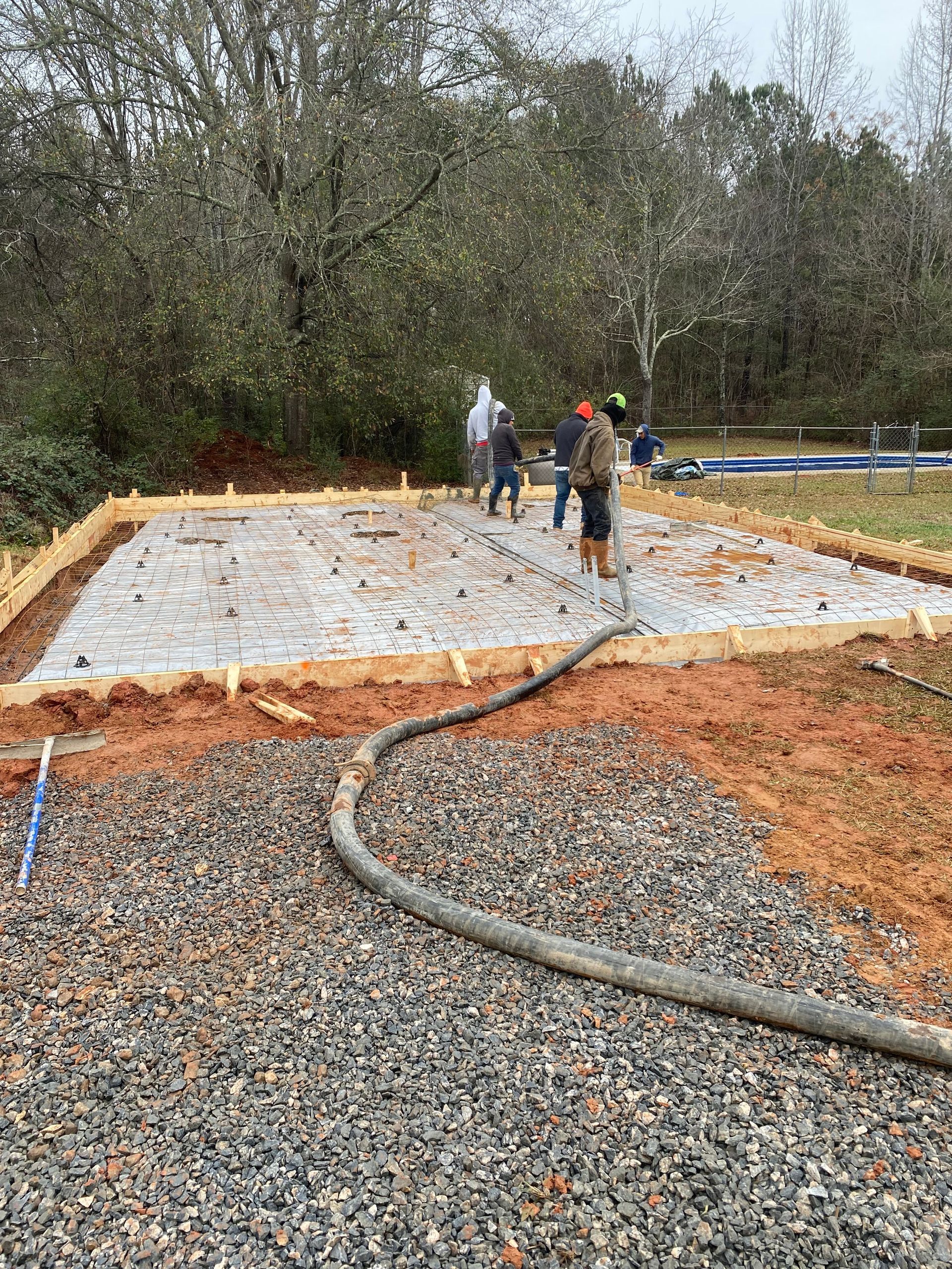Construction workers pouring concrete slab on a gravel base, using a hose; wooded background.