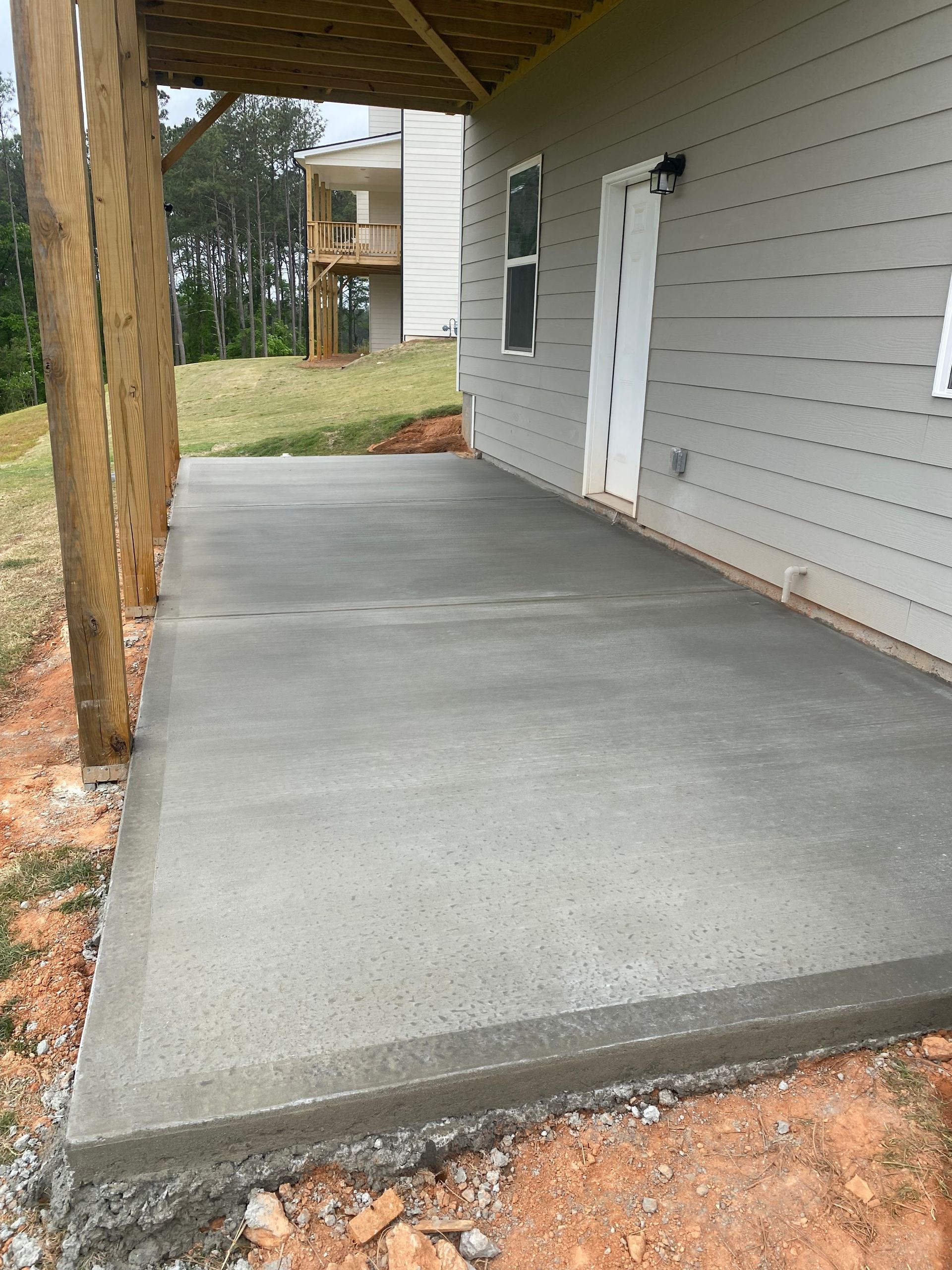 A newly poured concrete patio outside a house, with a wooden overhang and door.