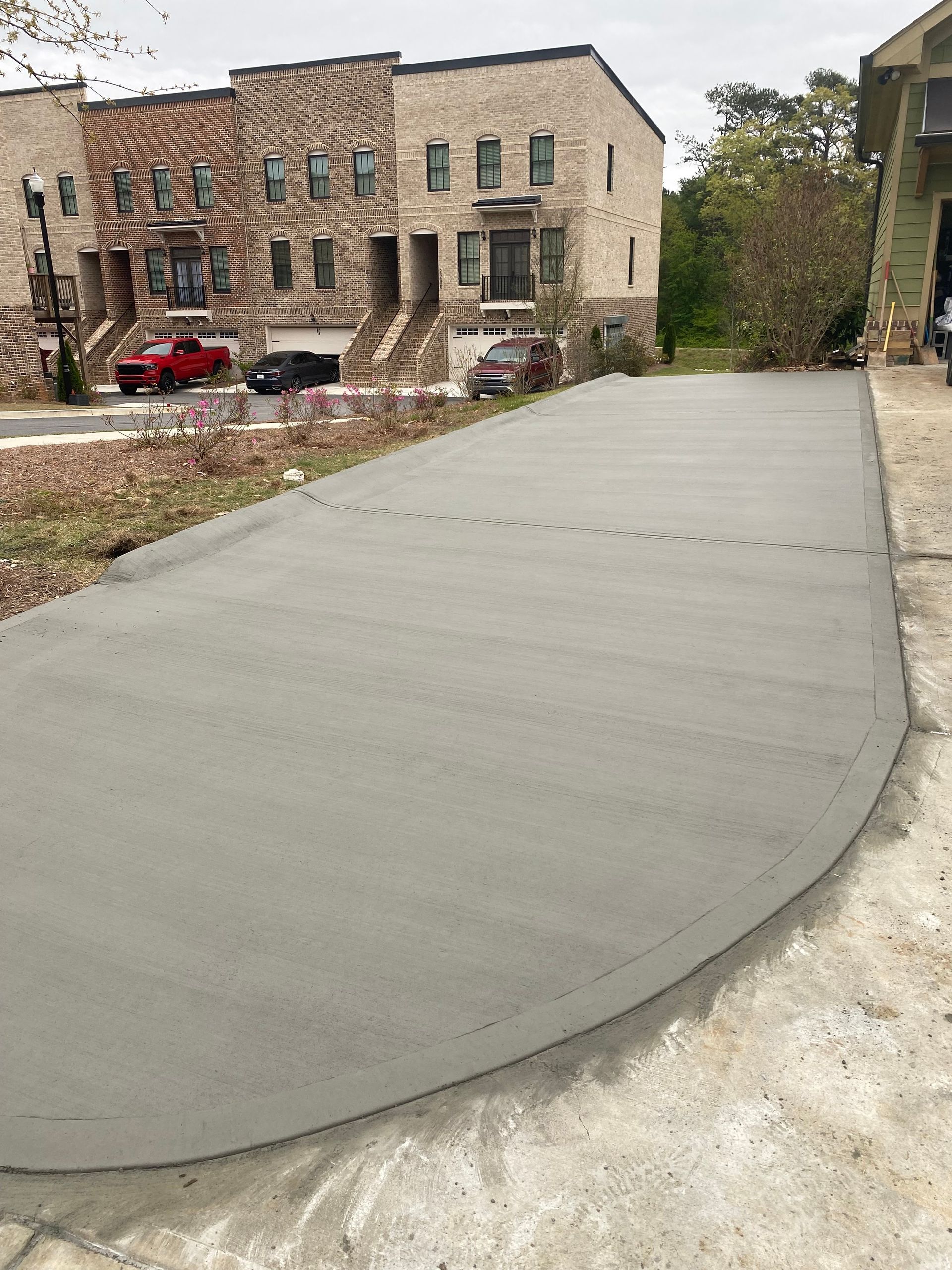 Newly poured concrete driveway with a rounded edge, leading to a street with parked cars and townhouses.
