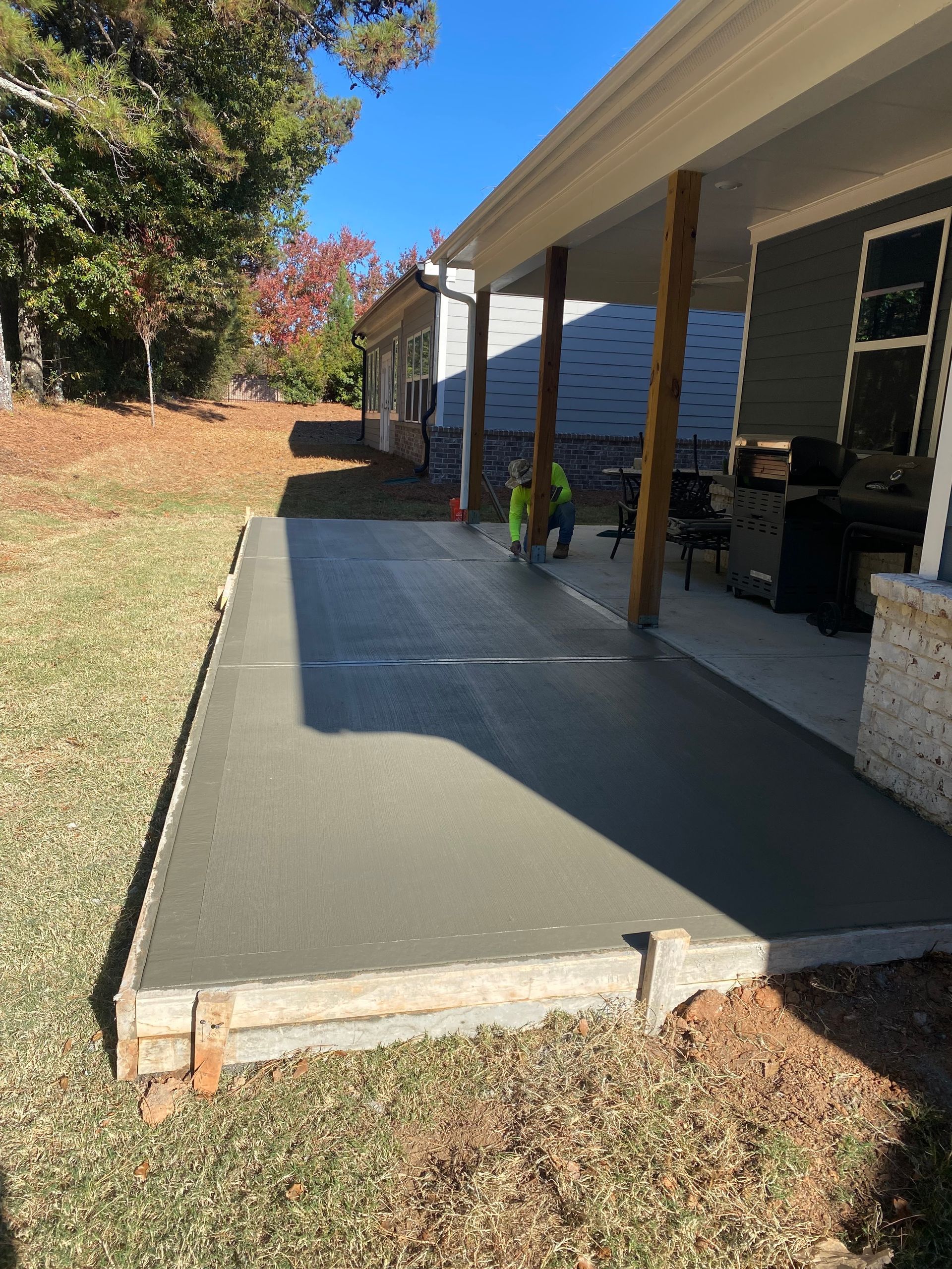 Newly poured concrete patio next to a house with a covered porch. Two workers in neon vests smooth the surface.