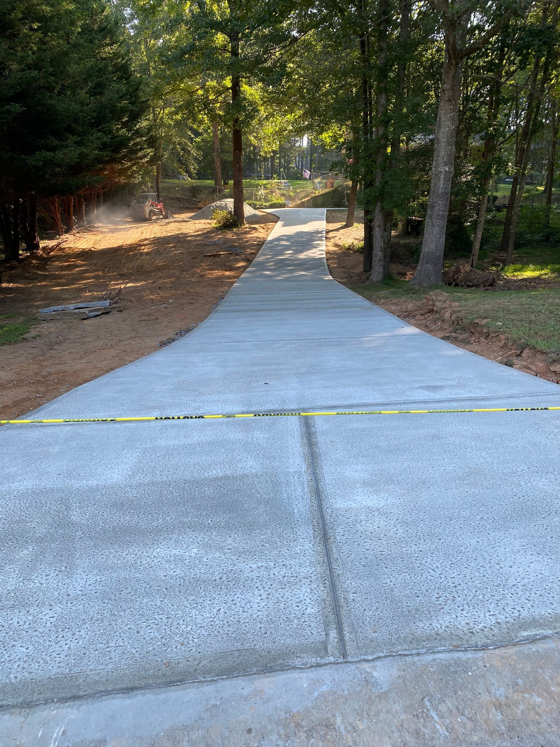 Newly poured concrete driveway, yellow caution tape across the foreground, trees on either side, sunlit path.