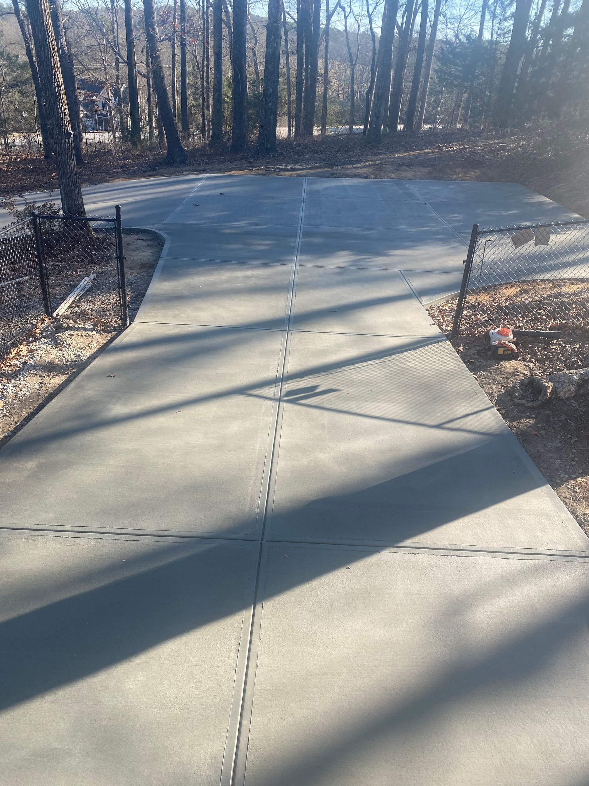 Concrete driveway leading to a wooded area, with joint lines and fencing on both sides.
