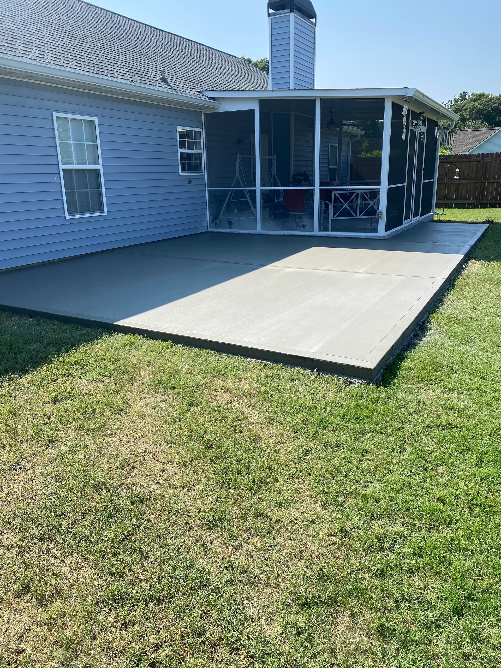 A new concrete patio with a screened porch attached to a blue house, in a grassy backyard.