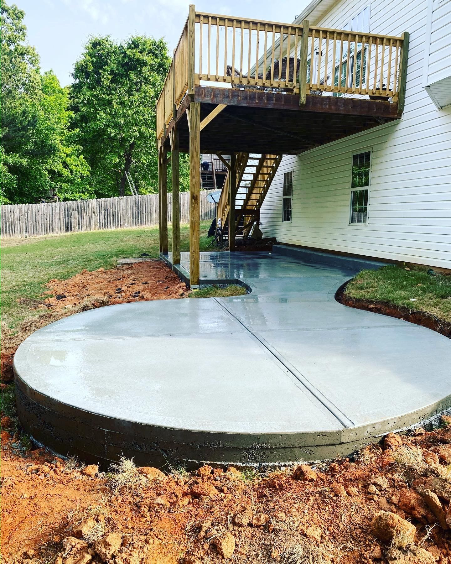 Newly poured concrete patio under a wooden deck attached to a house; dirt and grass surround.