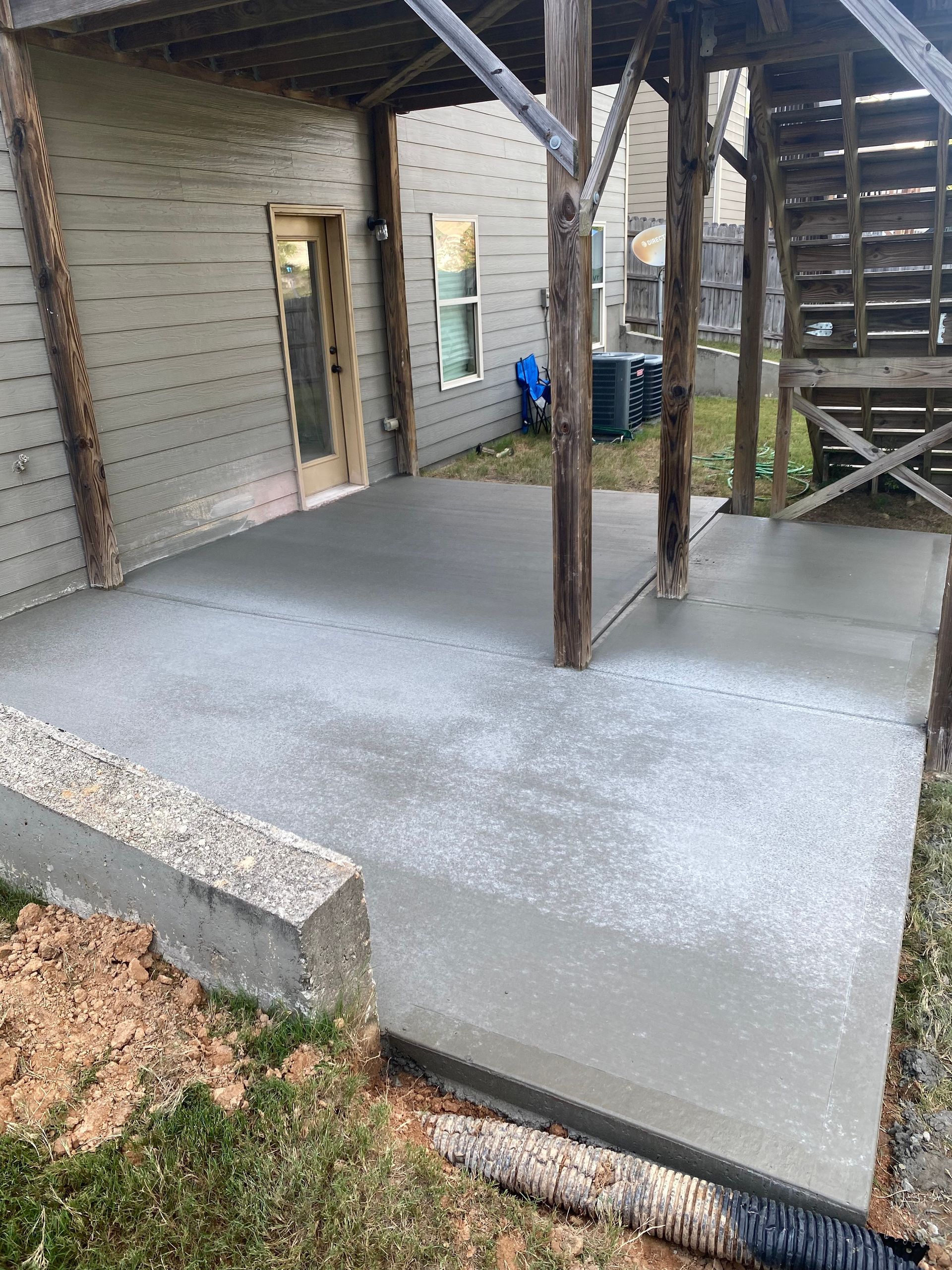 A freshly poured concrete patio next to a house with wooden supports and stairs, and a small garden.