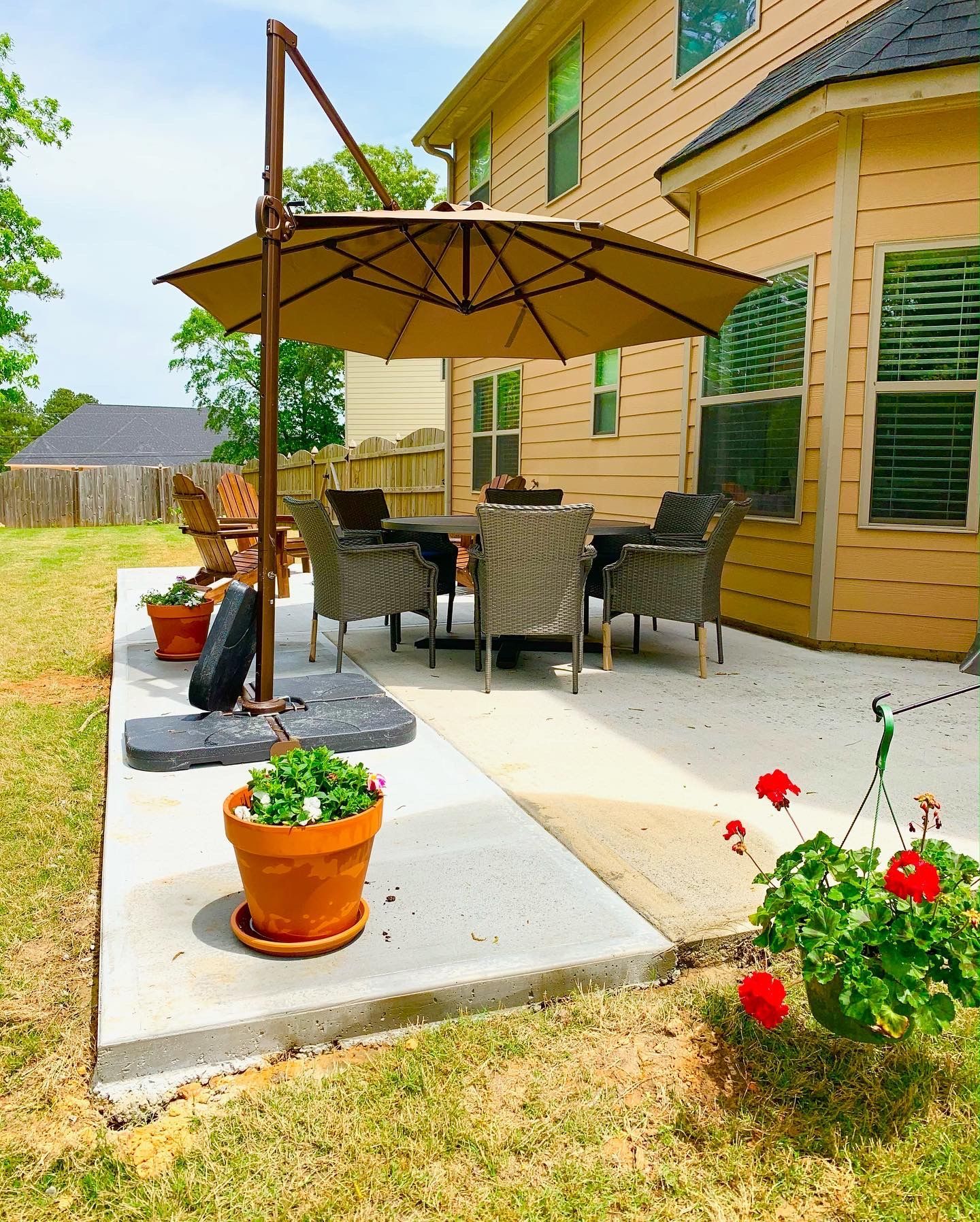 Patio with table, chairs, and umbrella. Pots with flowers. Beige house in the background.