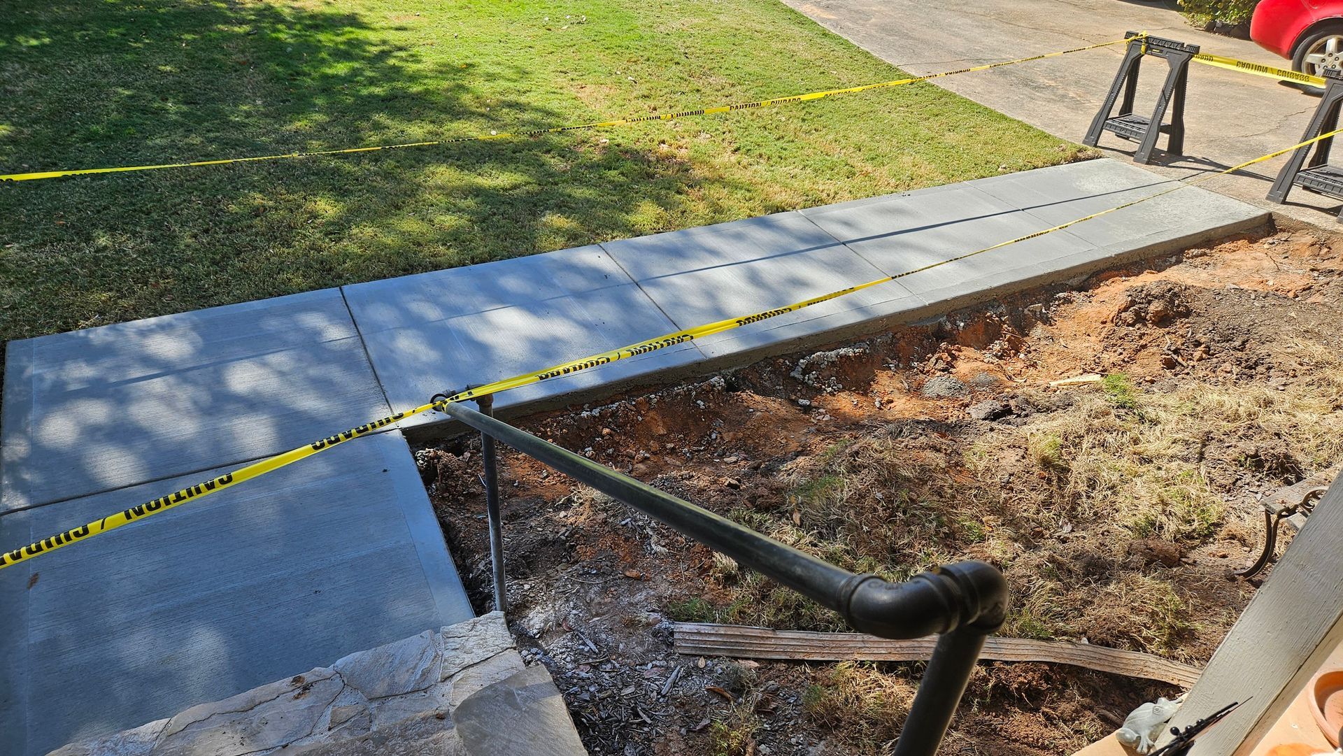 New concrete sidewalk under construction, blocked by yellow caution tape. Black railing, dirt and grass visible.