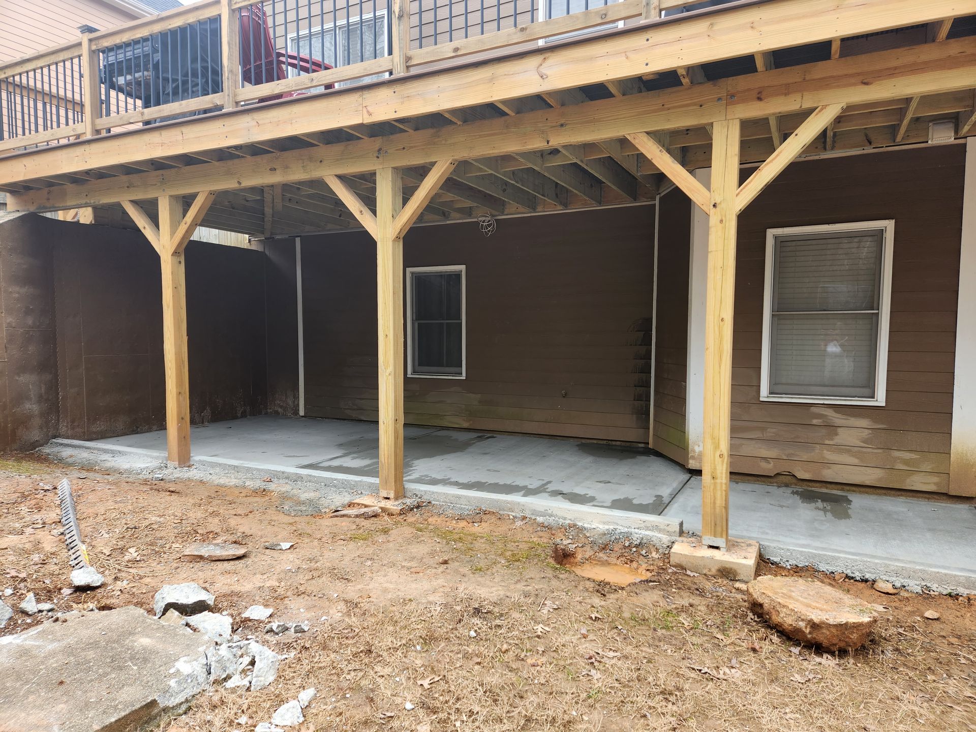 Deck supported by wooden posts over a concrete patio; brown exterior walls, two windows.