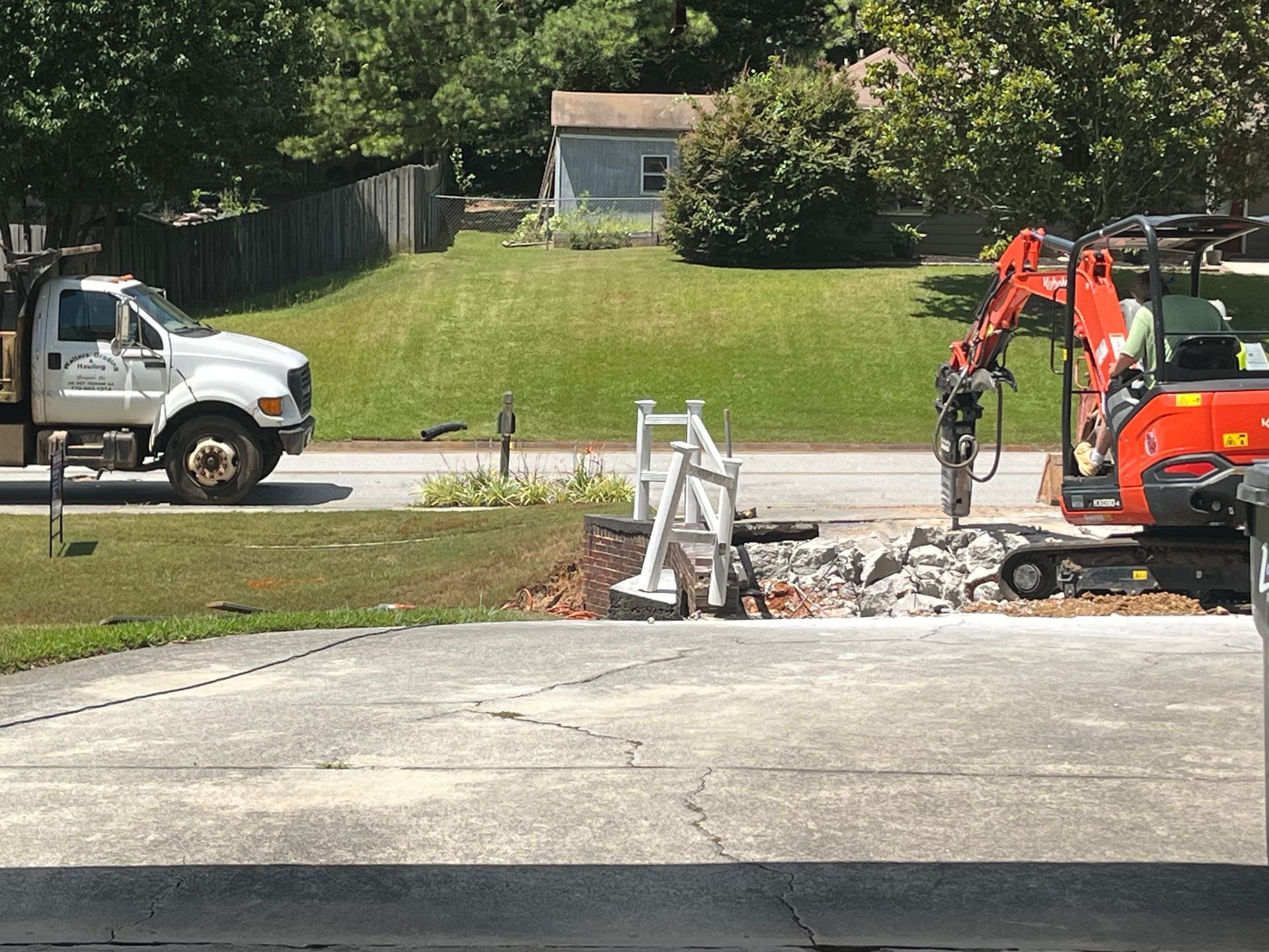 A small excavator breaking concrete in a driveway; a white truck is parked nearby.