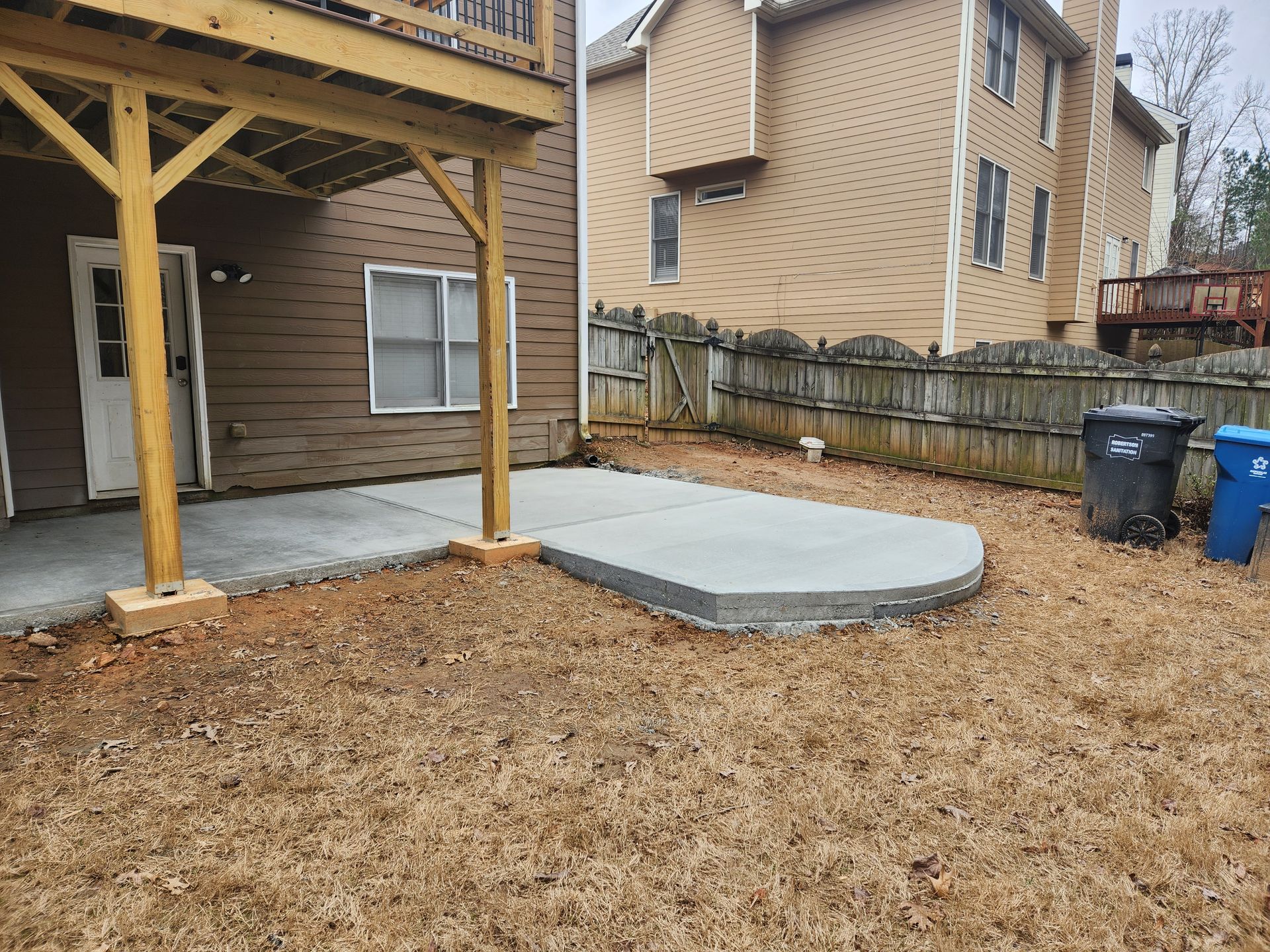A concrete patio extends from a house, with a curved edge and wooden deck above, surrounded by a yard with brown leaves.
