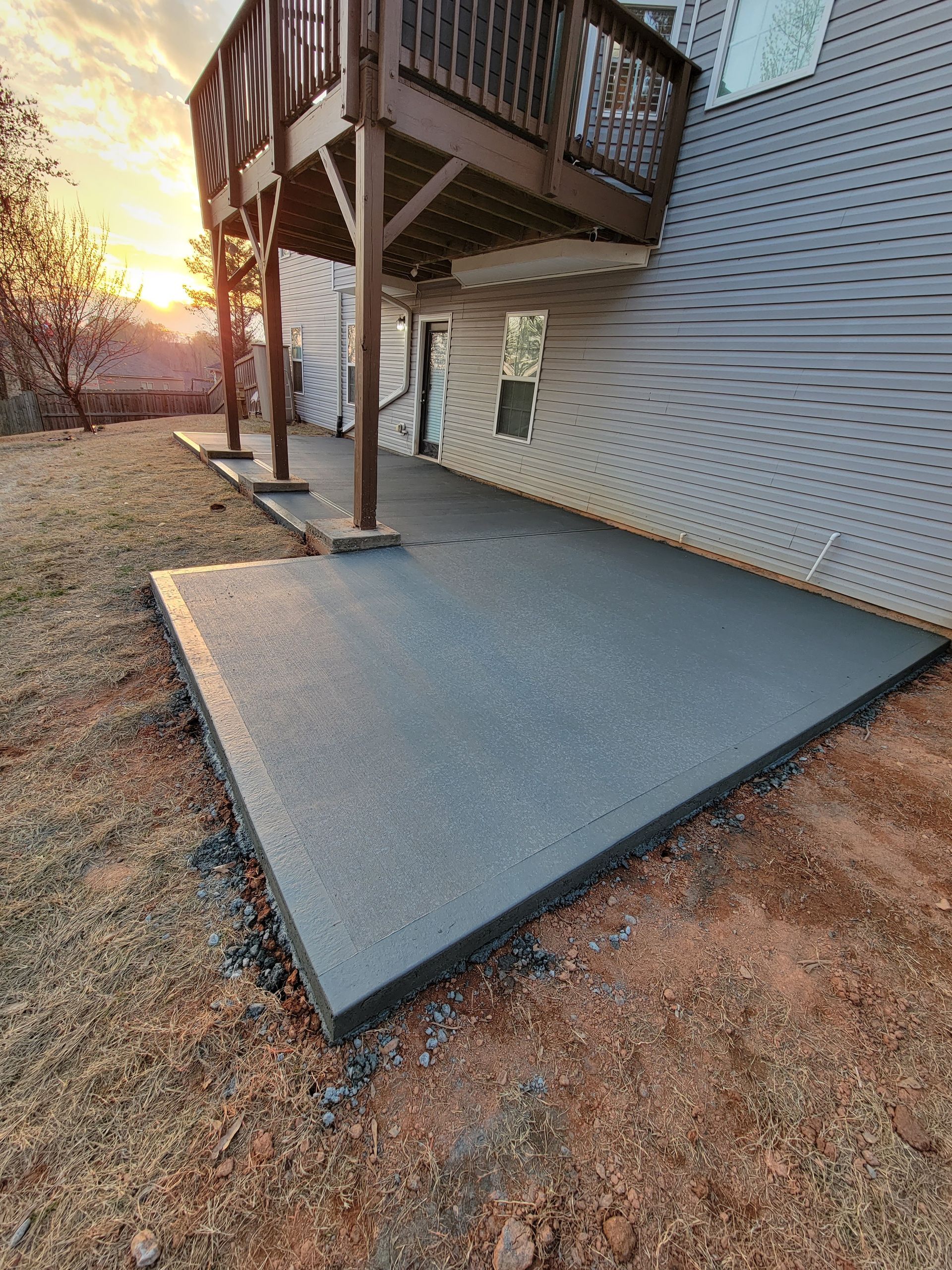 Gray concrete patio beneath a deck, set against the side of a house. Sunlight in the distance.