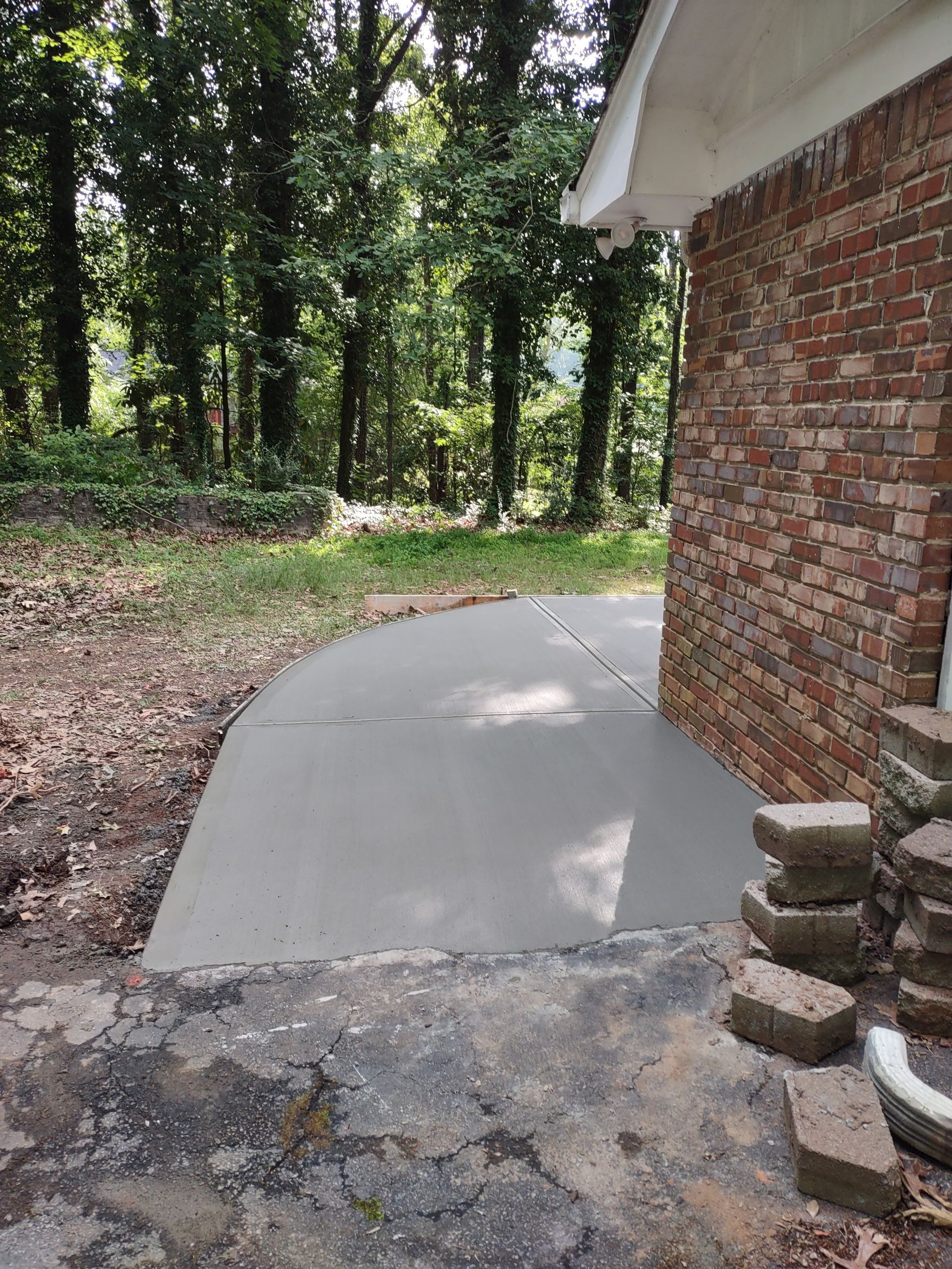 Newly poured concrete patio next to a brick building. Trees and foliage in the background.