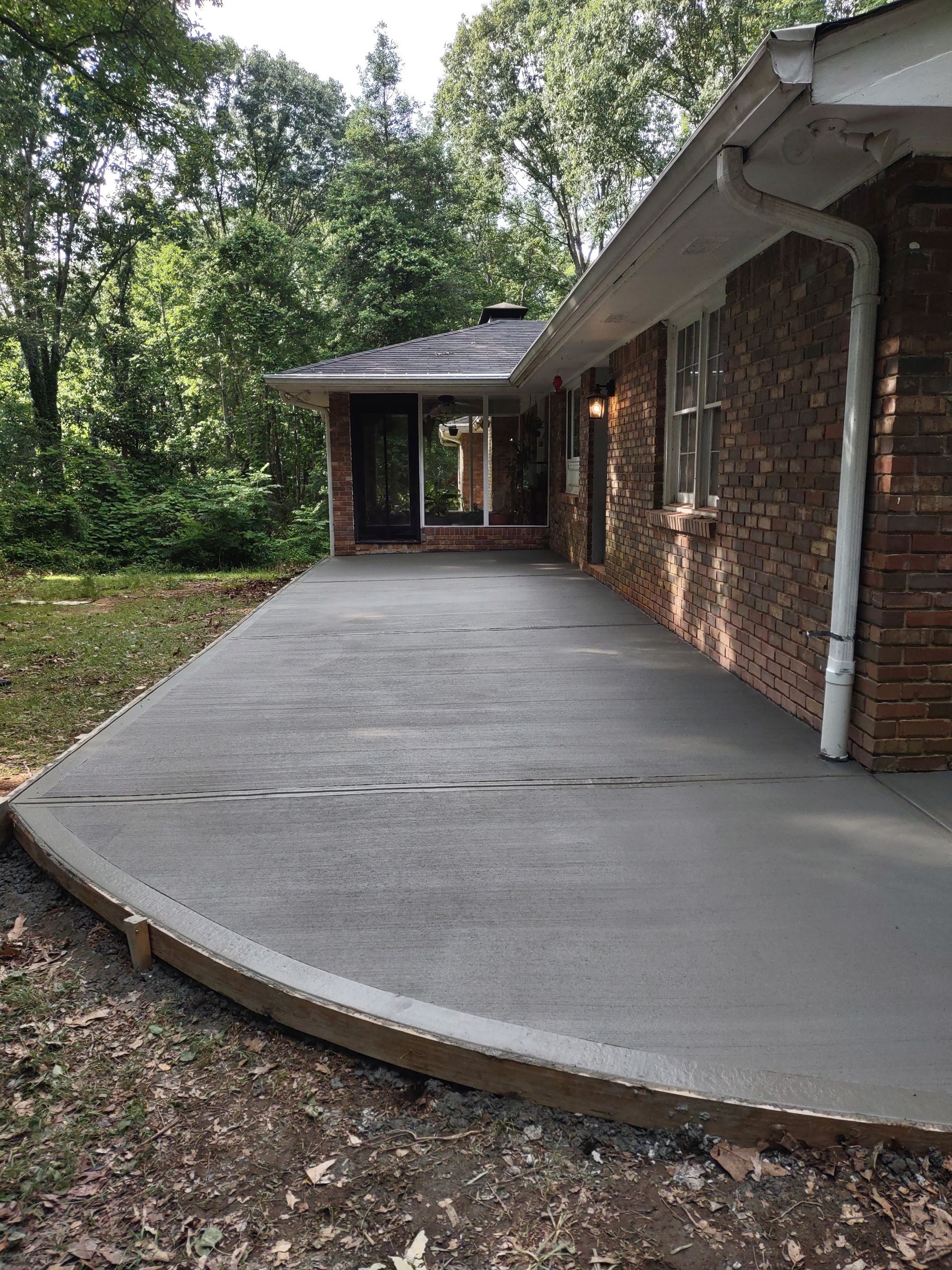 Newly poured concrete patio next to a brick house. Trees in the background.