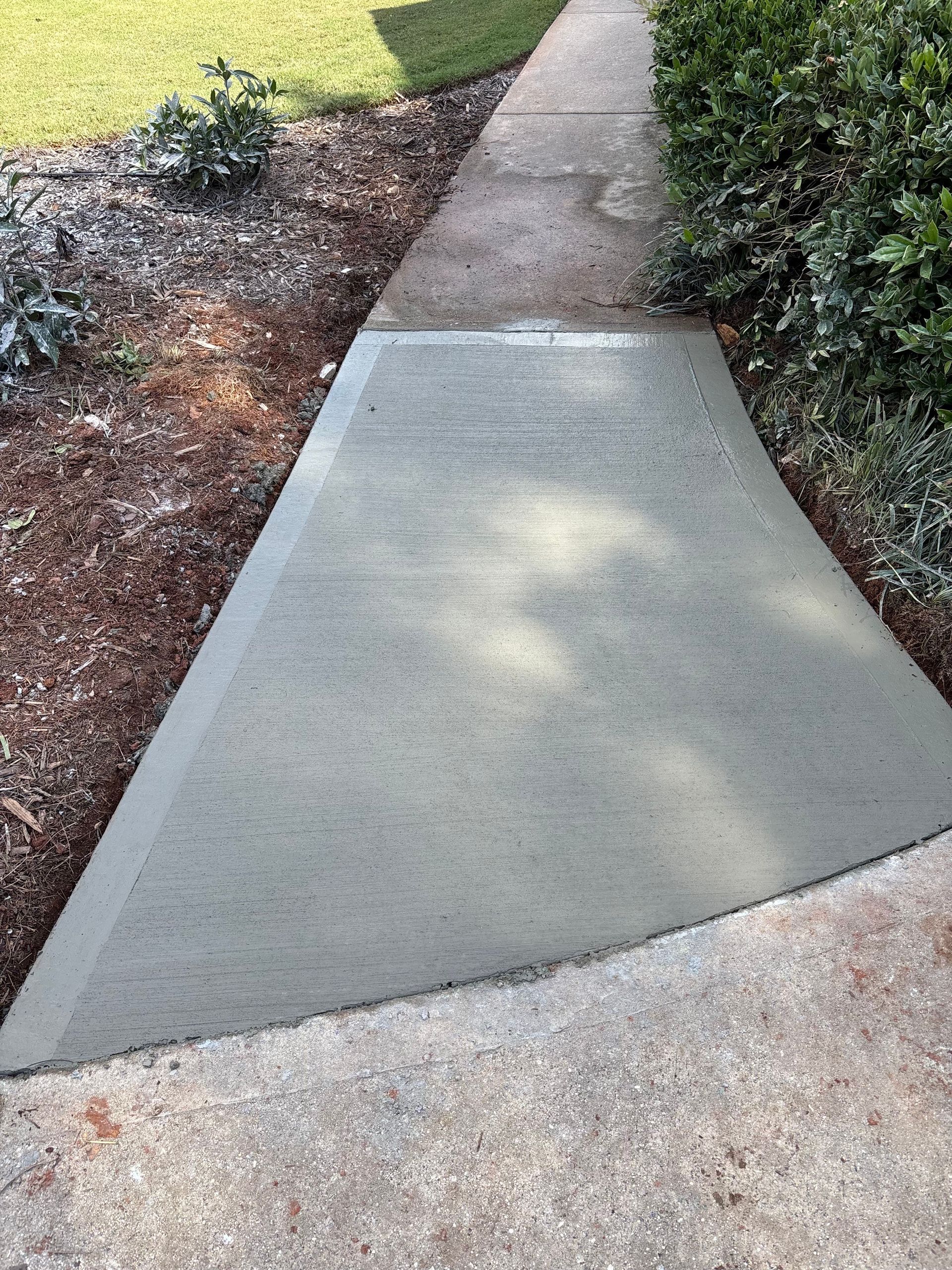 Freshly poured concrete sidewalk widening to a triangular shape, adjacent to existing walkway and landscaping.