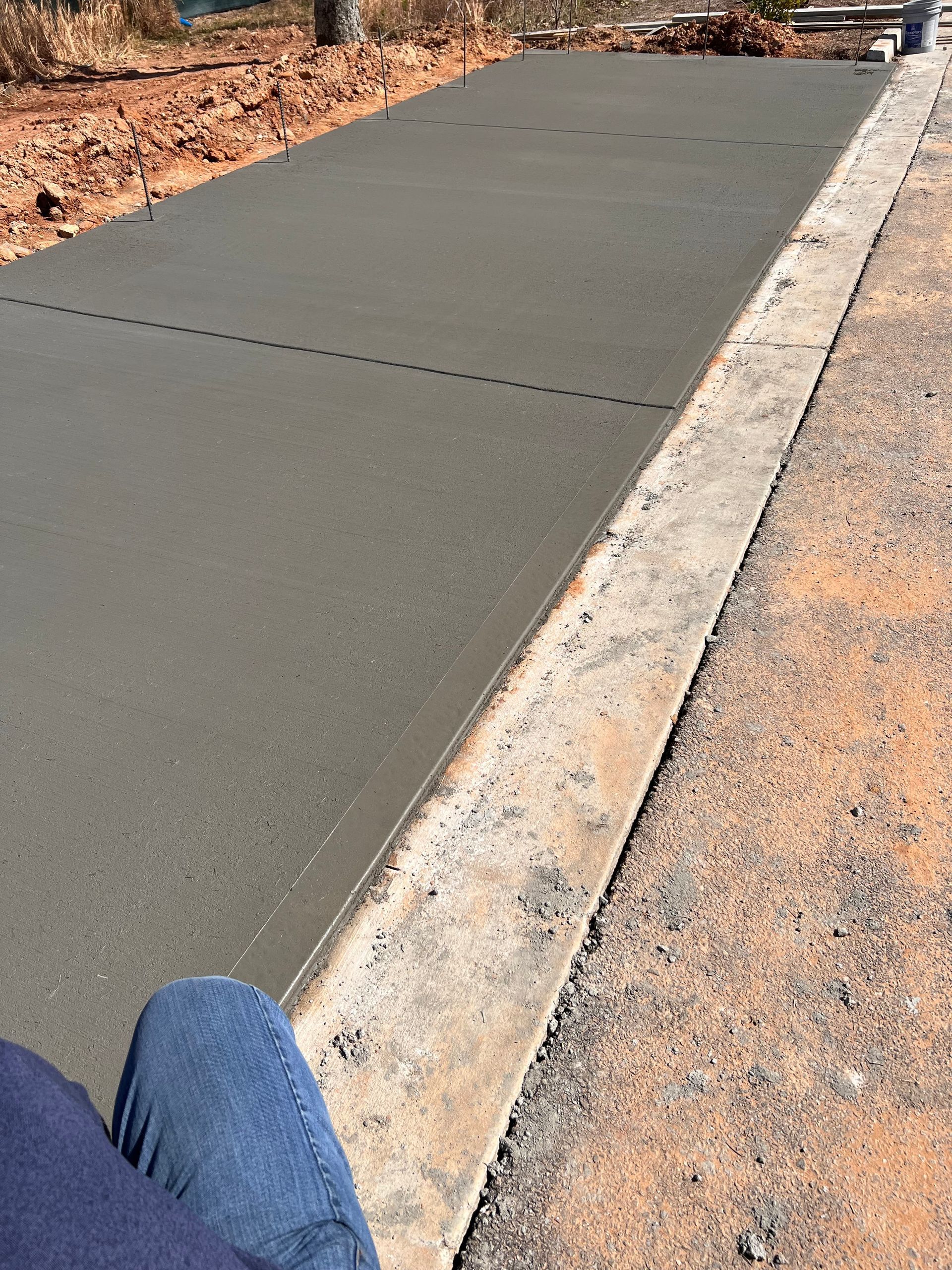 Freshly poured concrete slab with a dashed line, next to a raised curb and dirt.