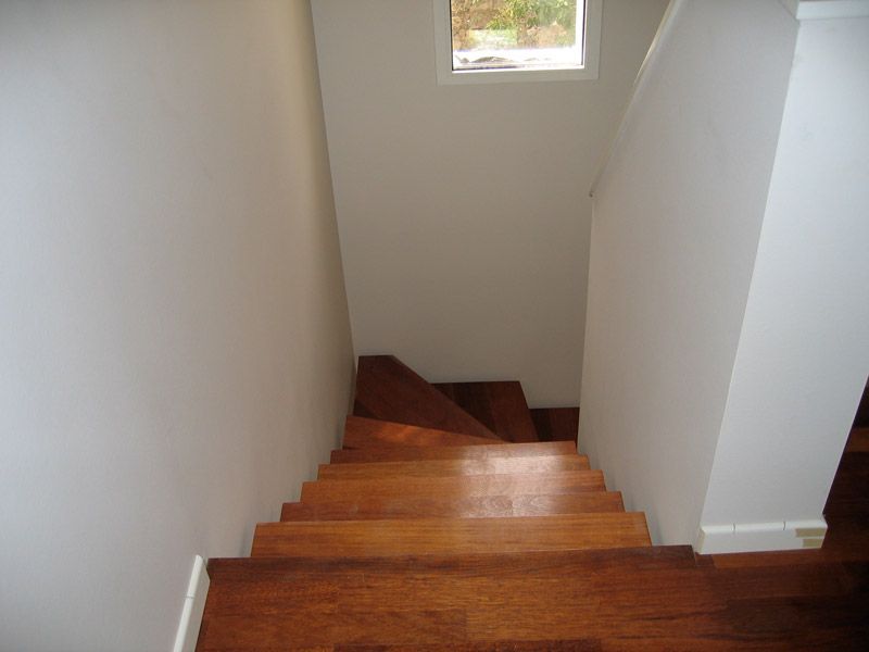 Escalier en bois descendant entre des murs blancs, avec une petite fenêtre en haut.