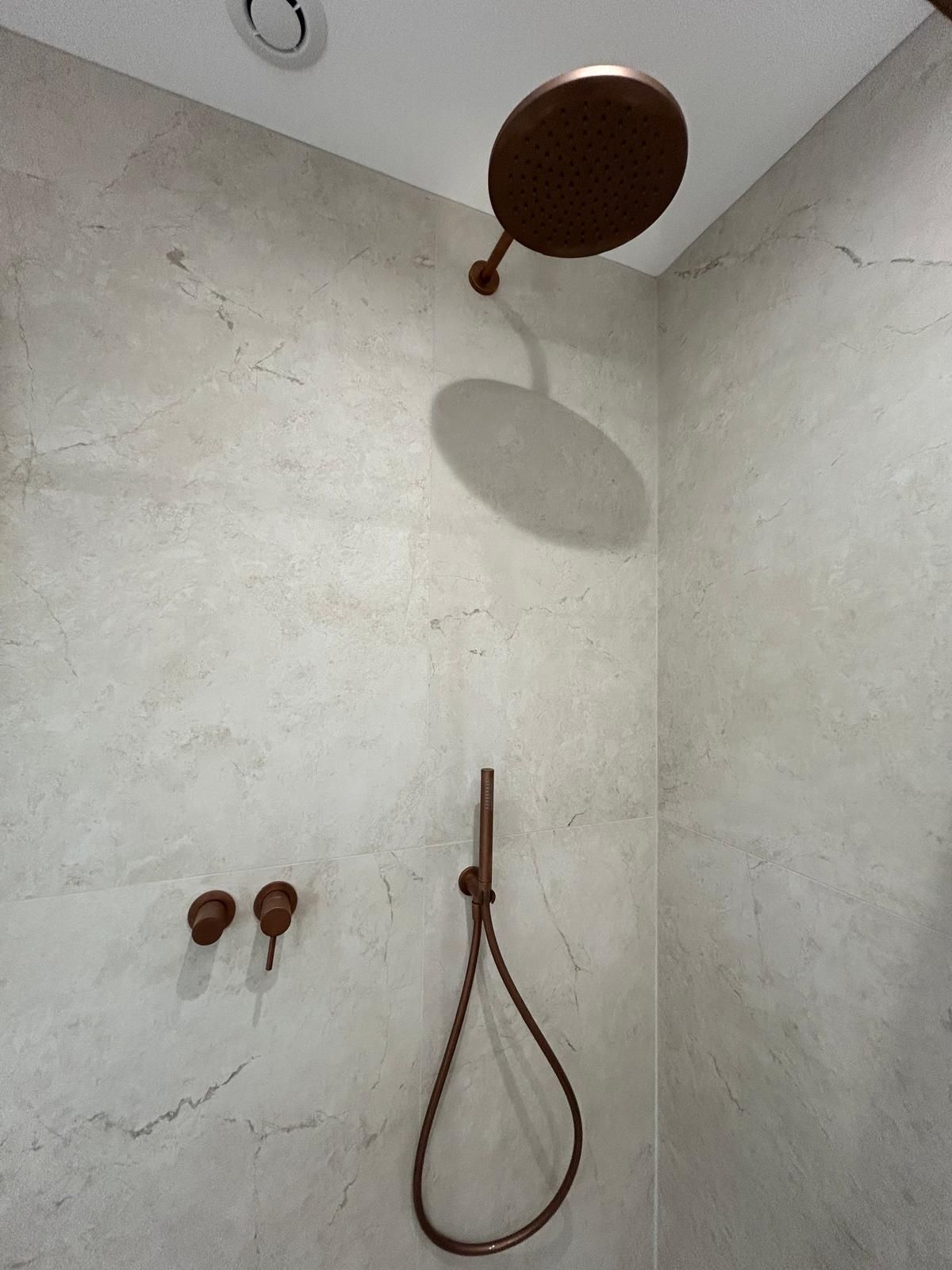 Pomme de douche et accessoires en bronze sur carrelage de couleur claire dans une douche.
