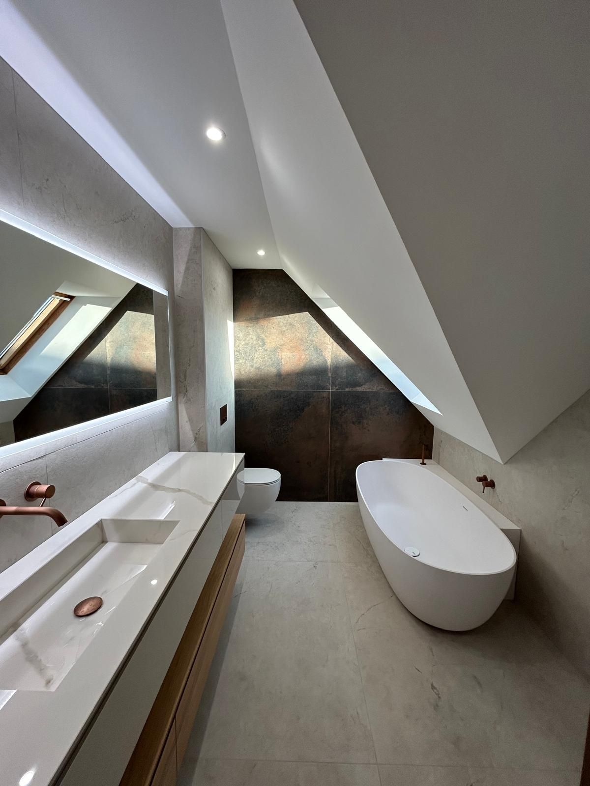 Salle de bain moderne avec plafond incliné; baignoire, lavabo et toilettes blancs; luminaires en cuivre; mur marron.