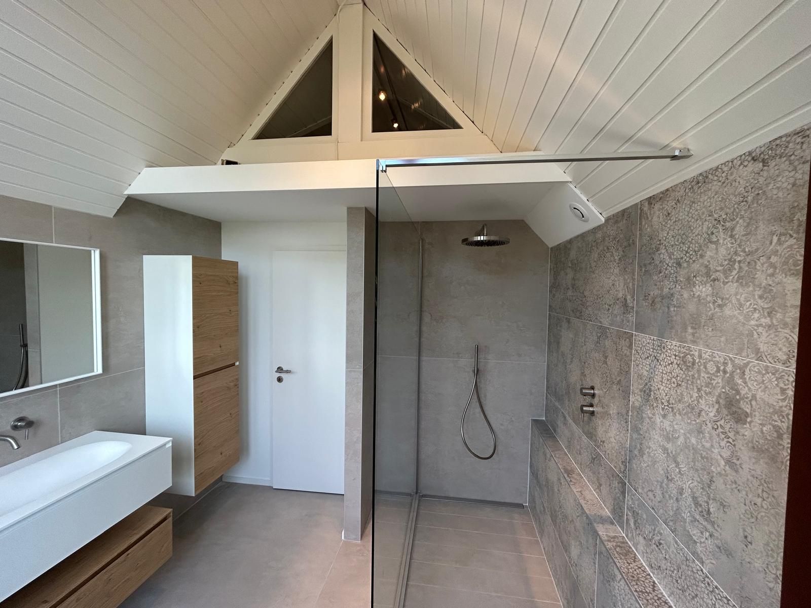Salle de bain moderne avec douche et lavabo. Tons neutres, carrelage gris, touches de bois et fenêtre triangulaire.