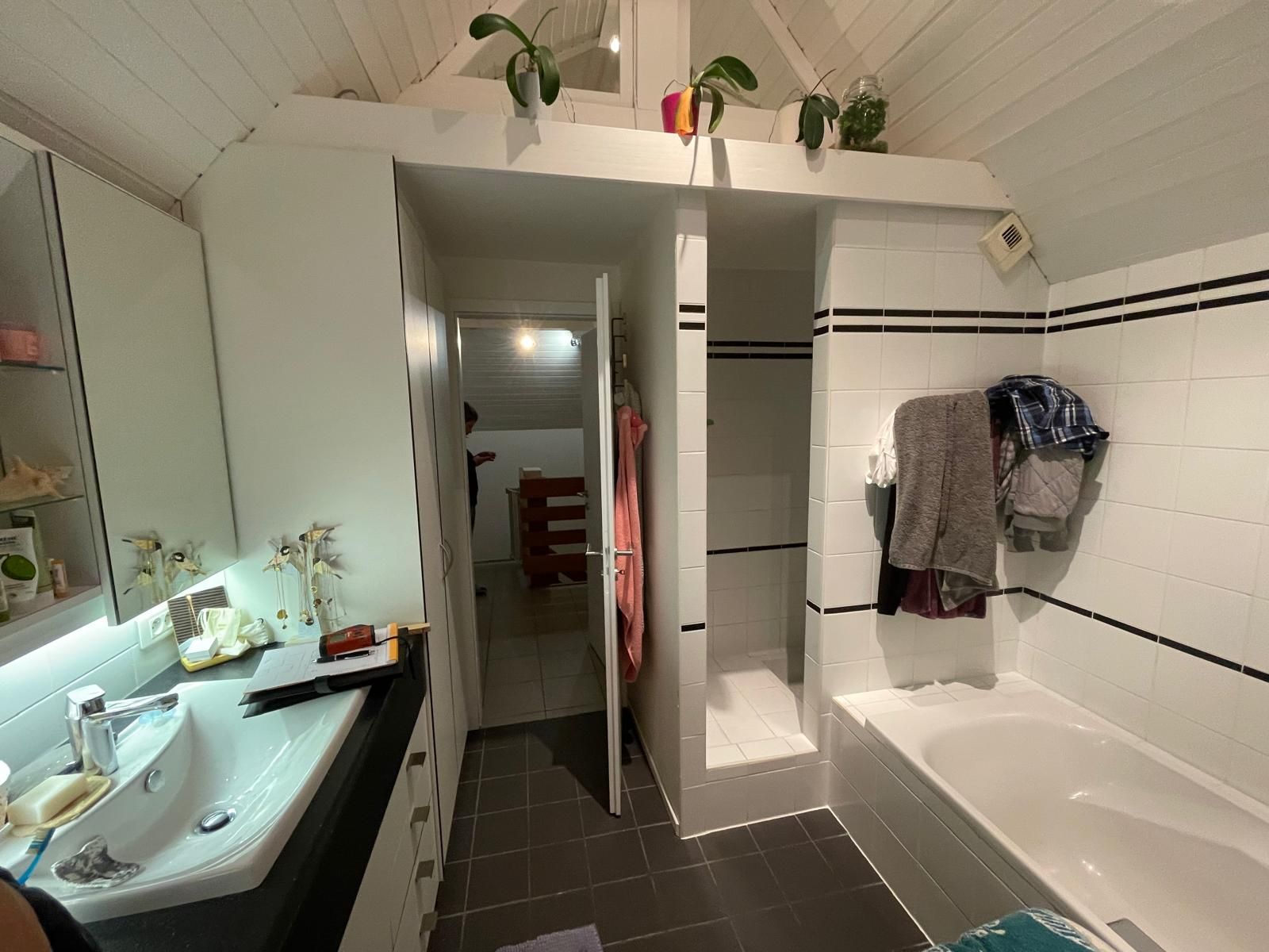 Salle de bain aux murs blancs, avec lavabo, baignoire et douche ouverte. Une porte mène à une autre pièce.