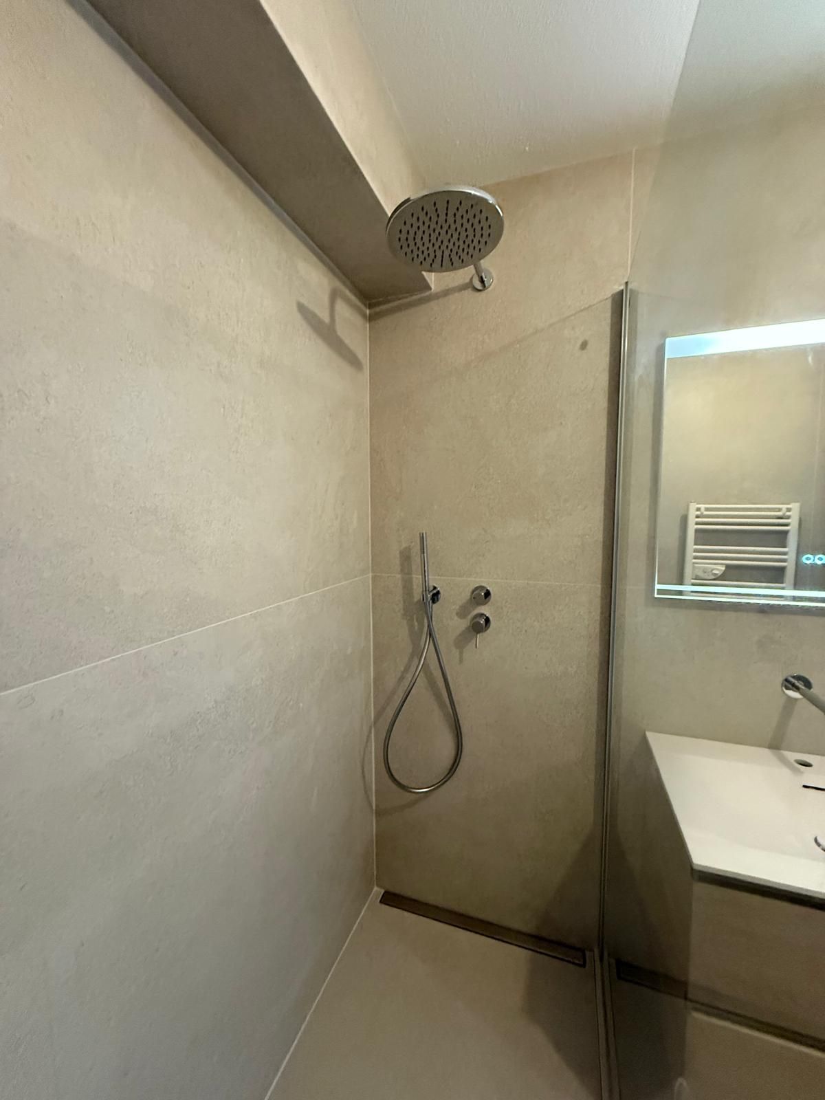 Cabine de douche avec murs gris, pommeau de douche à effet pluie, douche à main et porte vitrée.