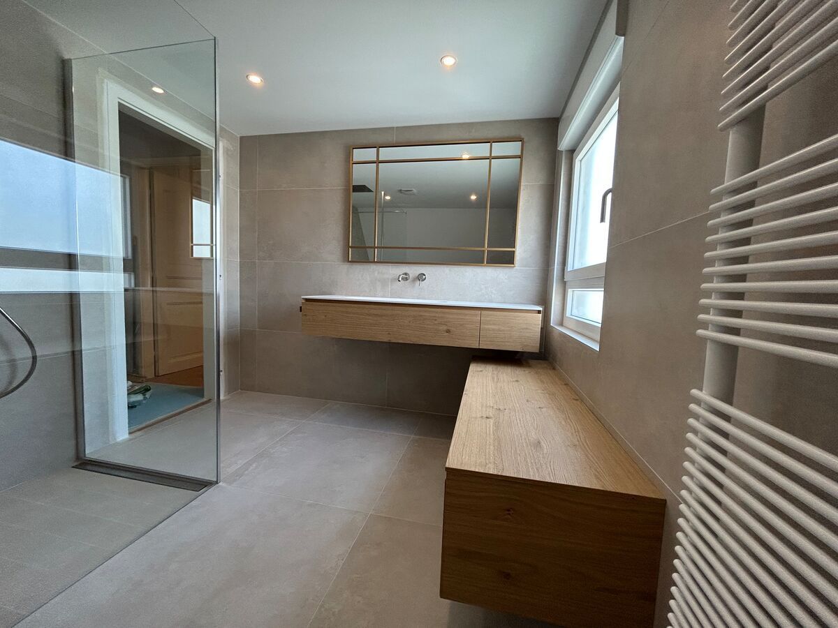 Salle de bain moderne avec vanité et banc en bois, douche en verre et murs et sol en carrelage de couleur neutre.